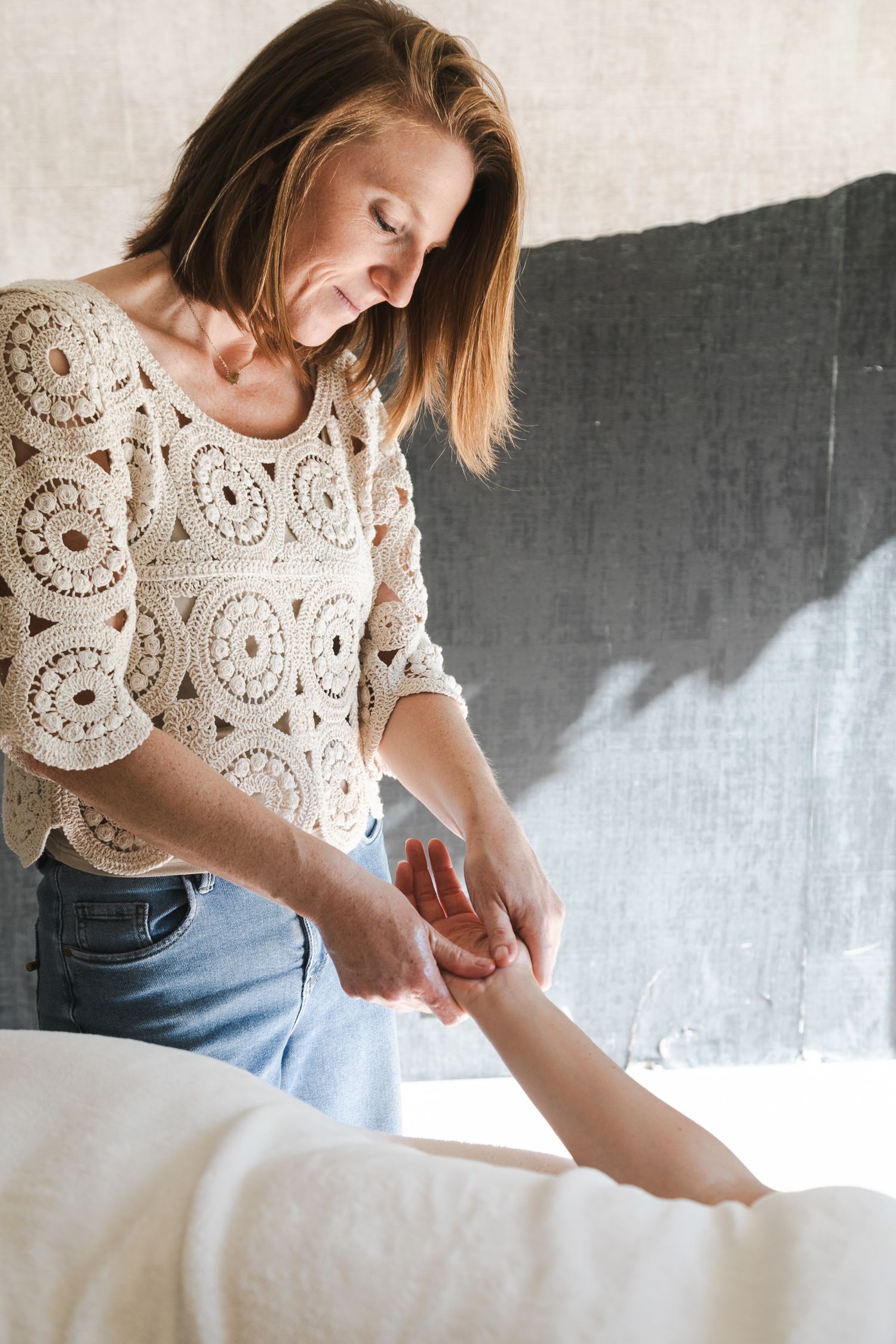 Praticienne douce, souriante, debout et à coté de sa cliente. Elle lui masse la main pour un moment de reconnexion après son accouchement.