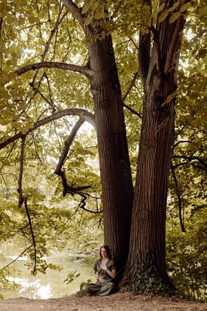 Praticienne en Reïki assise en tailleur au pied d'un grand arbre pour une méditation et un soin énergétique.
