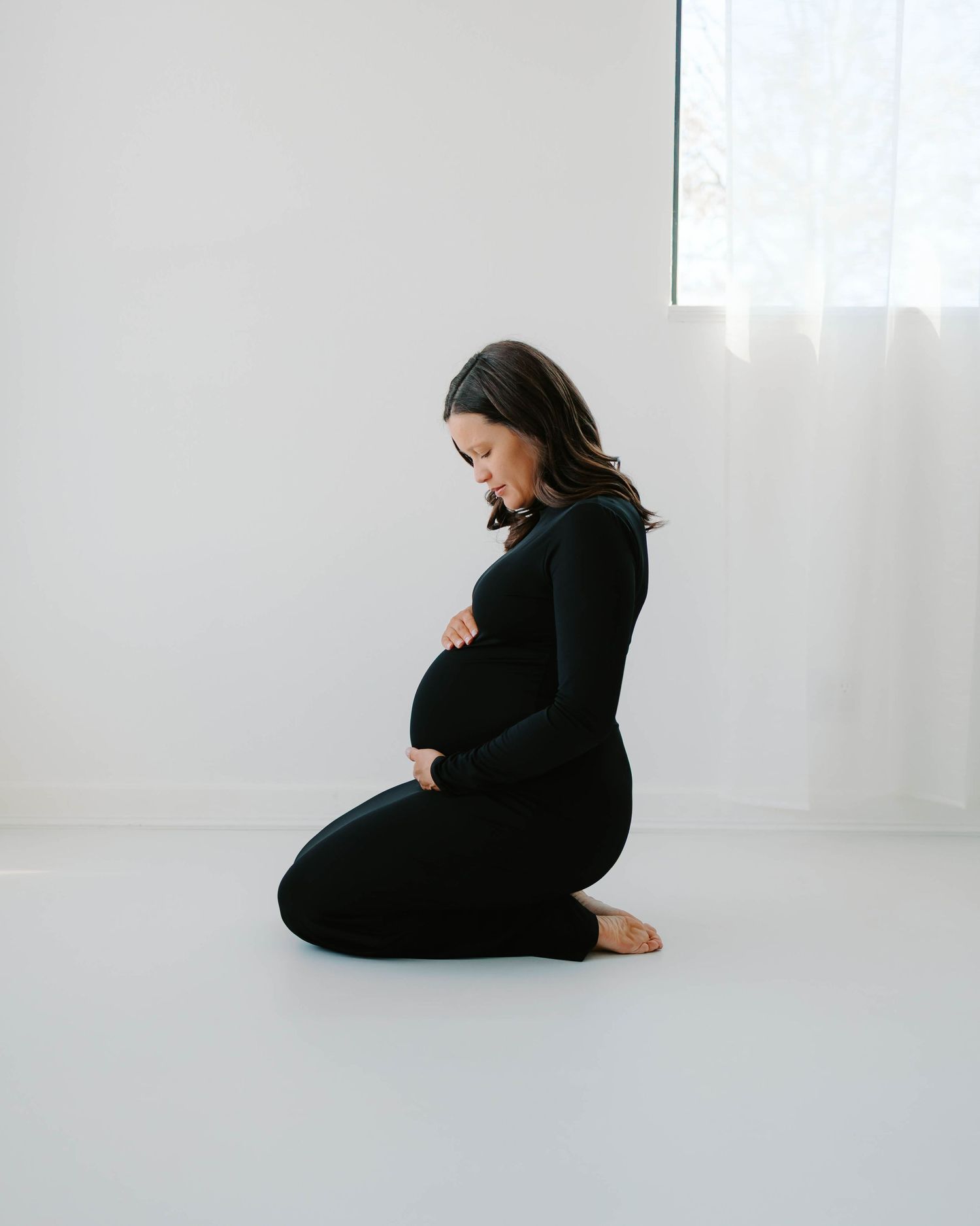 Pregnant woman in black dress sitting down holding her belly in natural light studio- St. Louis, Missouri maternity photography