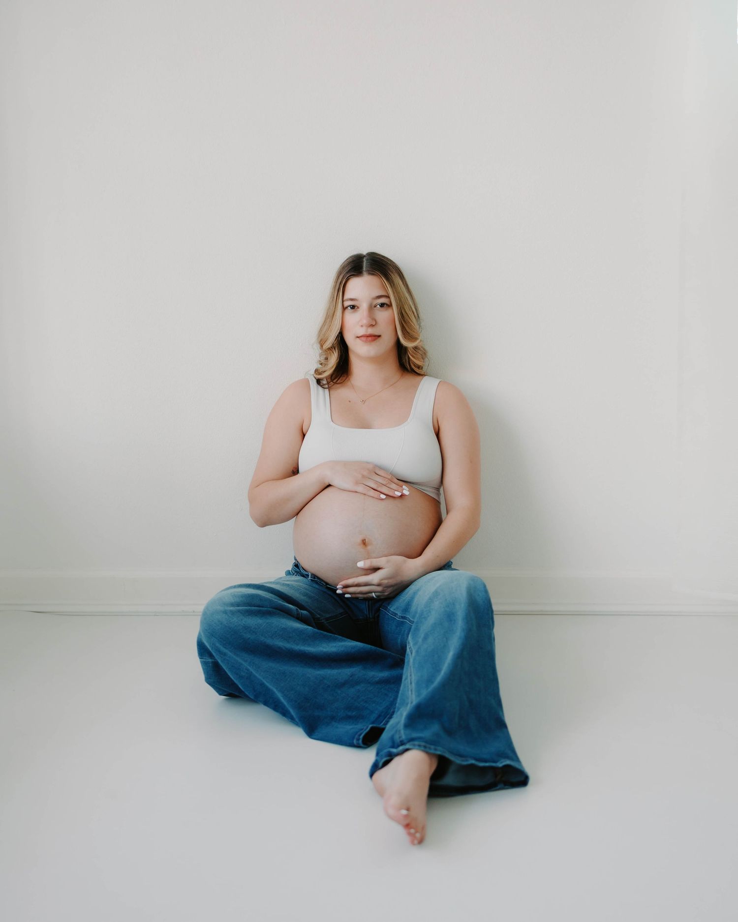 Pregnant mother in jeans and white shirt sitting down and holding her belly in natural light studio- St. Louis Missouri maternity session