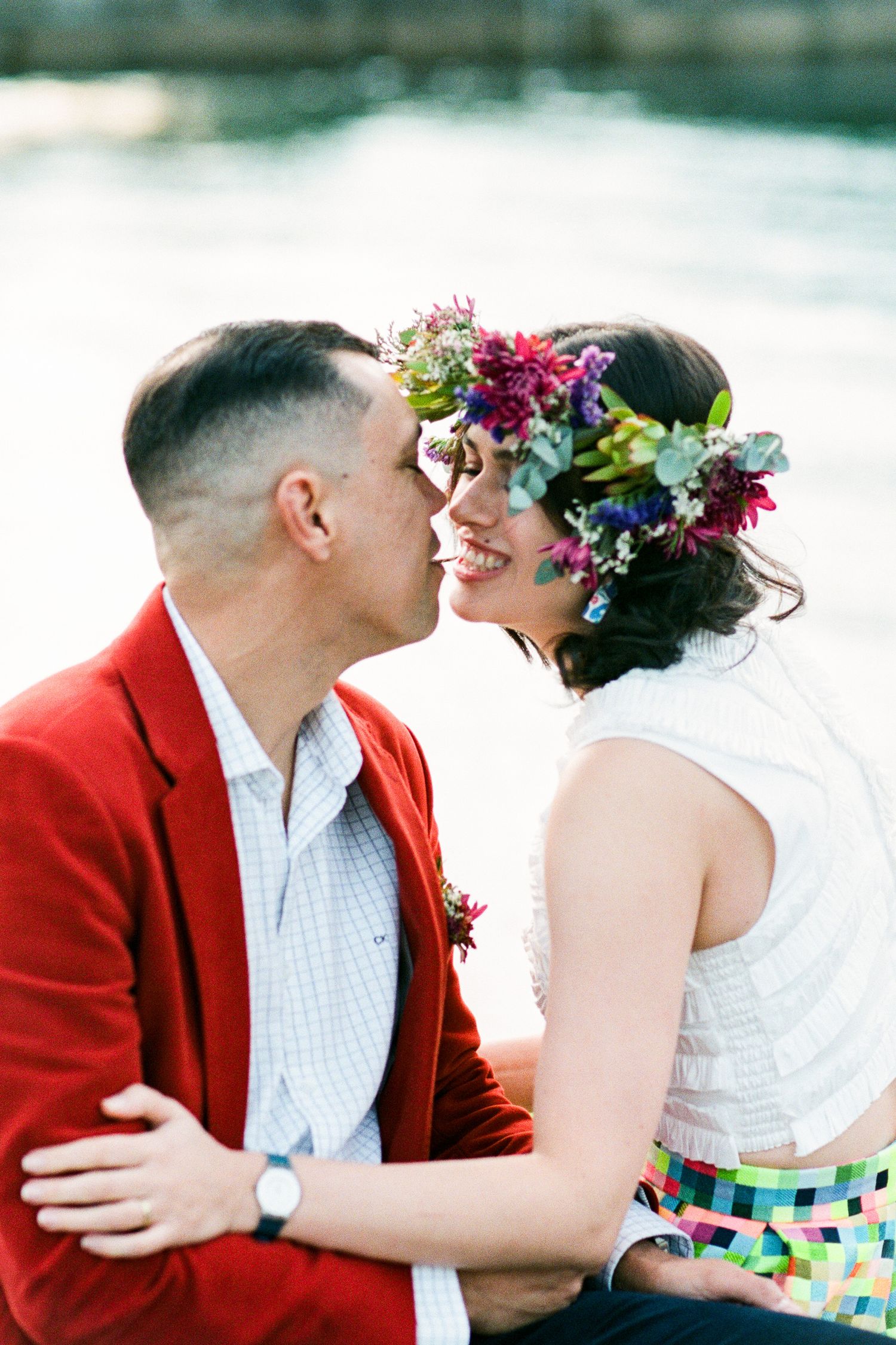 Couple in wedding attire sharing romantic moments by the water with colorful floral crown details.