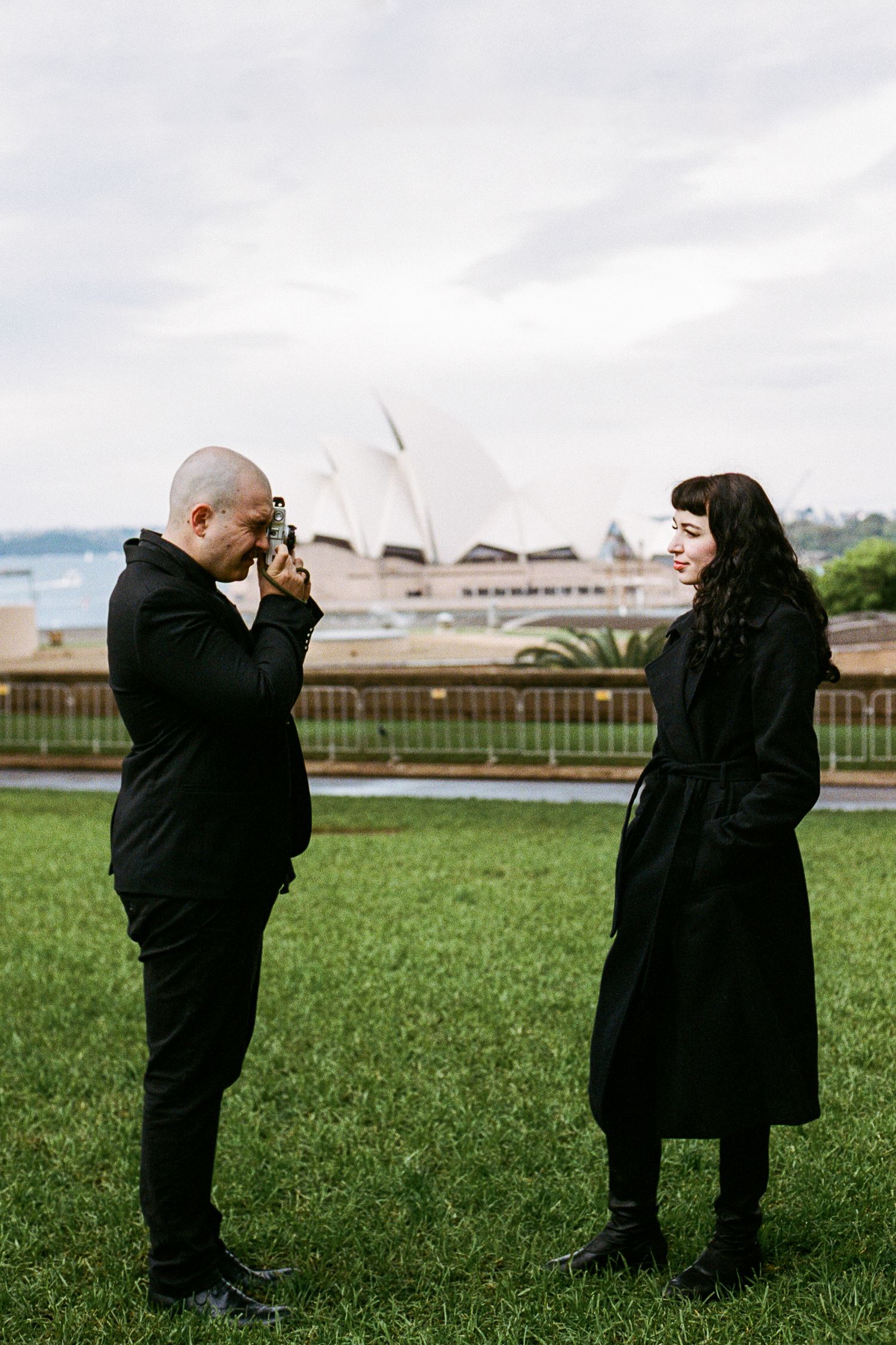Two people in black coats stand together with Sydney Opera House visible in background.