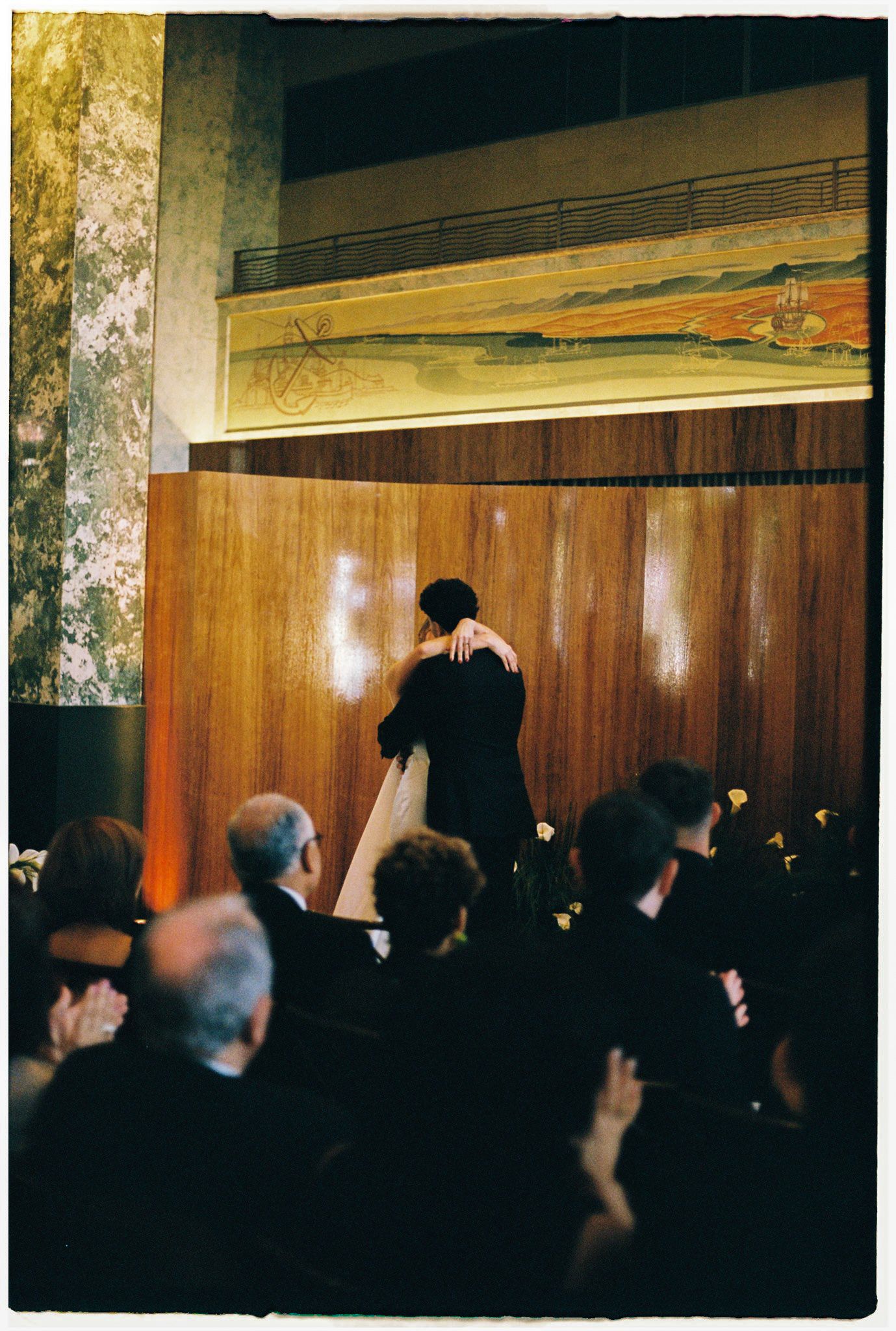 Performer bowing to applauding audience in elegant wood-paneled hall with mural above stage.