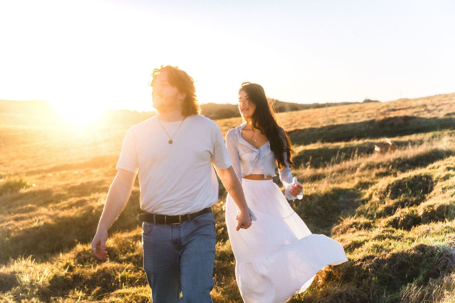 Couple walking hand in hand through a golden field at sunset while wearing white and denim outfits.