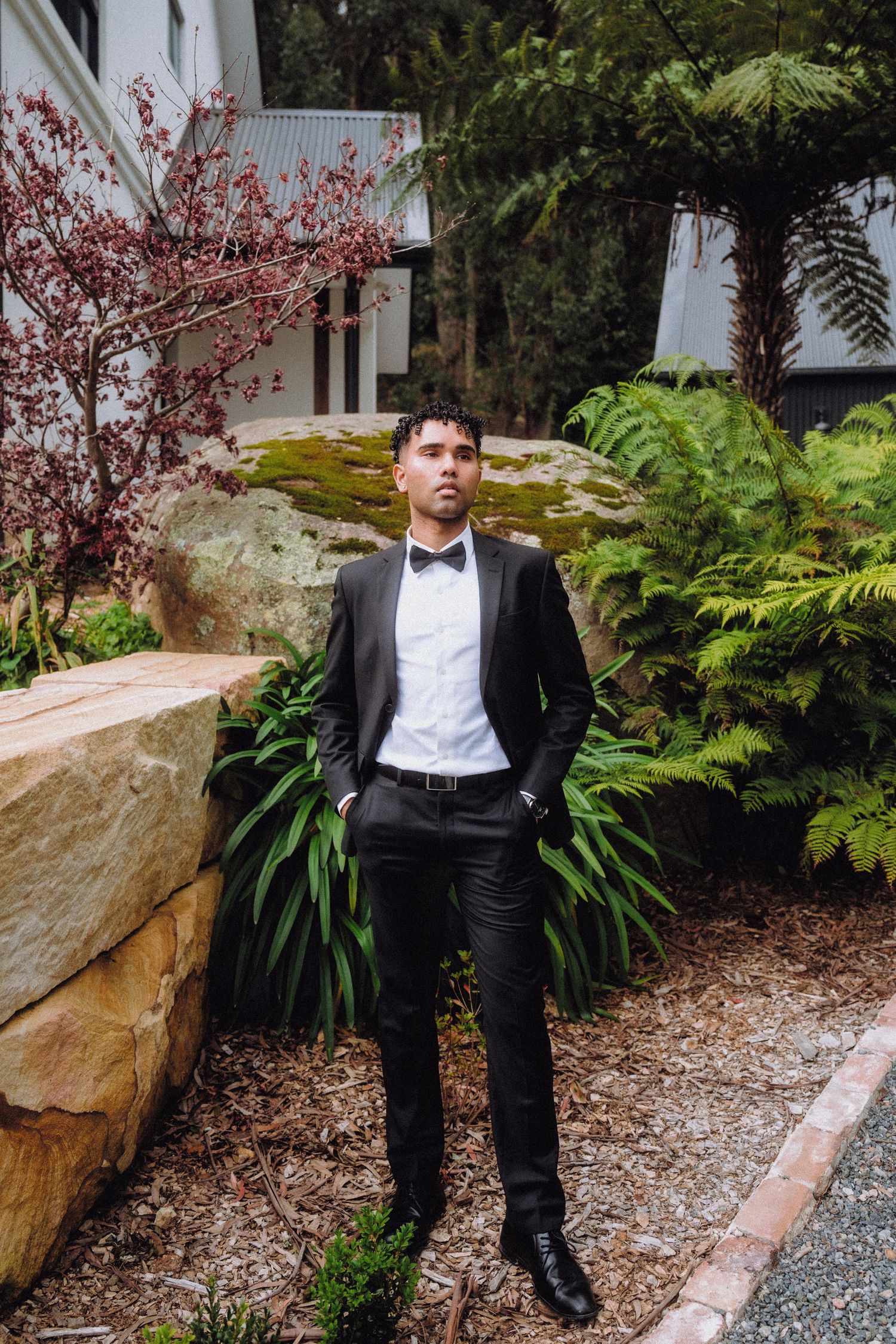 Dapper gentleman in bow tie poses against garden landscape with blooming red flowers.