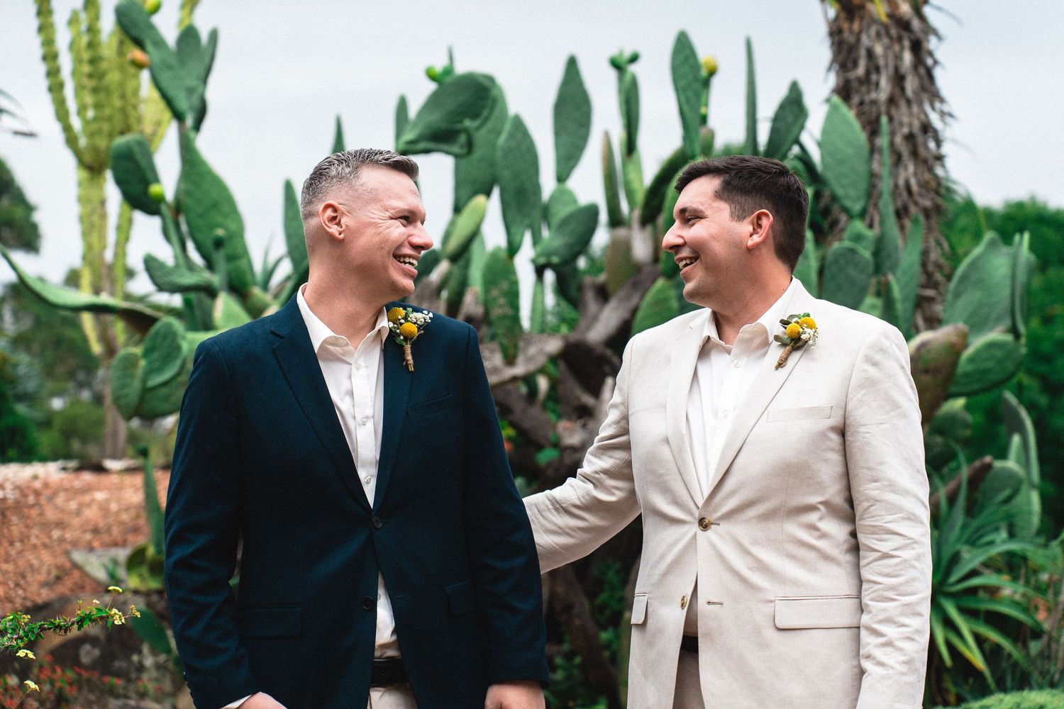 Two grooms in suits laughing together outdoors with cactus garden backdrop on their wedding day.