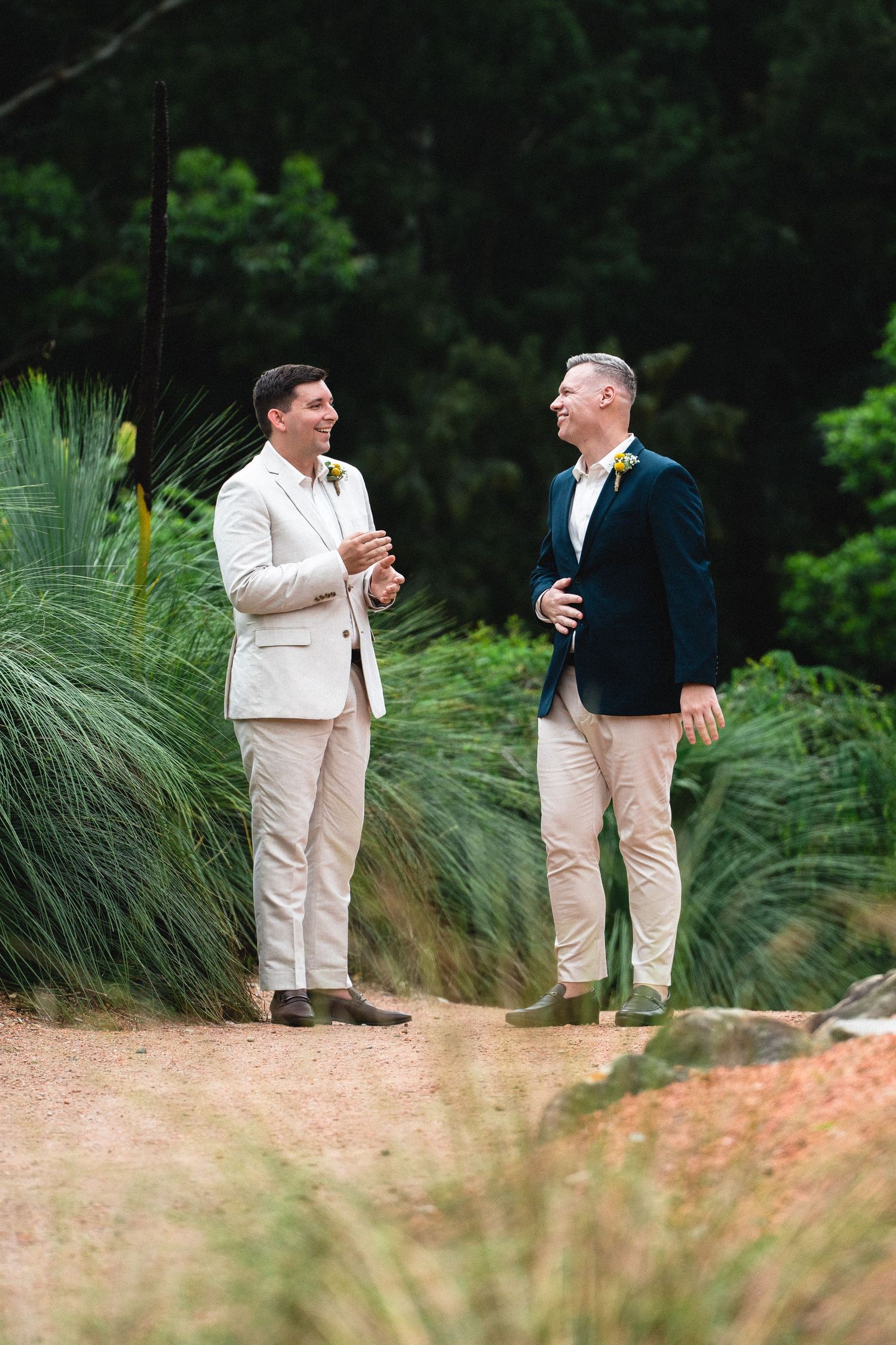 Two grooms in smart casual attire laugh together outdoors at a garden wedding ceremony.