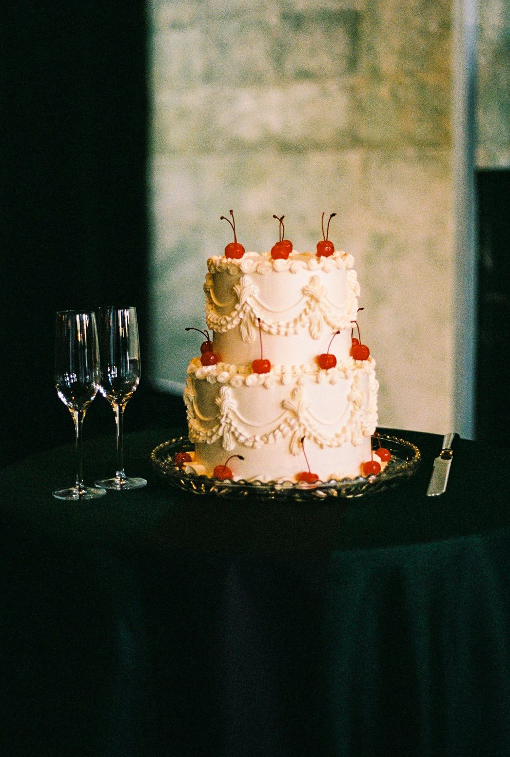 Elegant two-tier white wedding cake decorated with red cherries and pearl details beside champagne glasses.