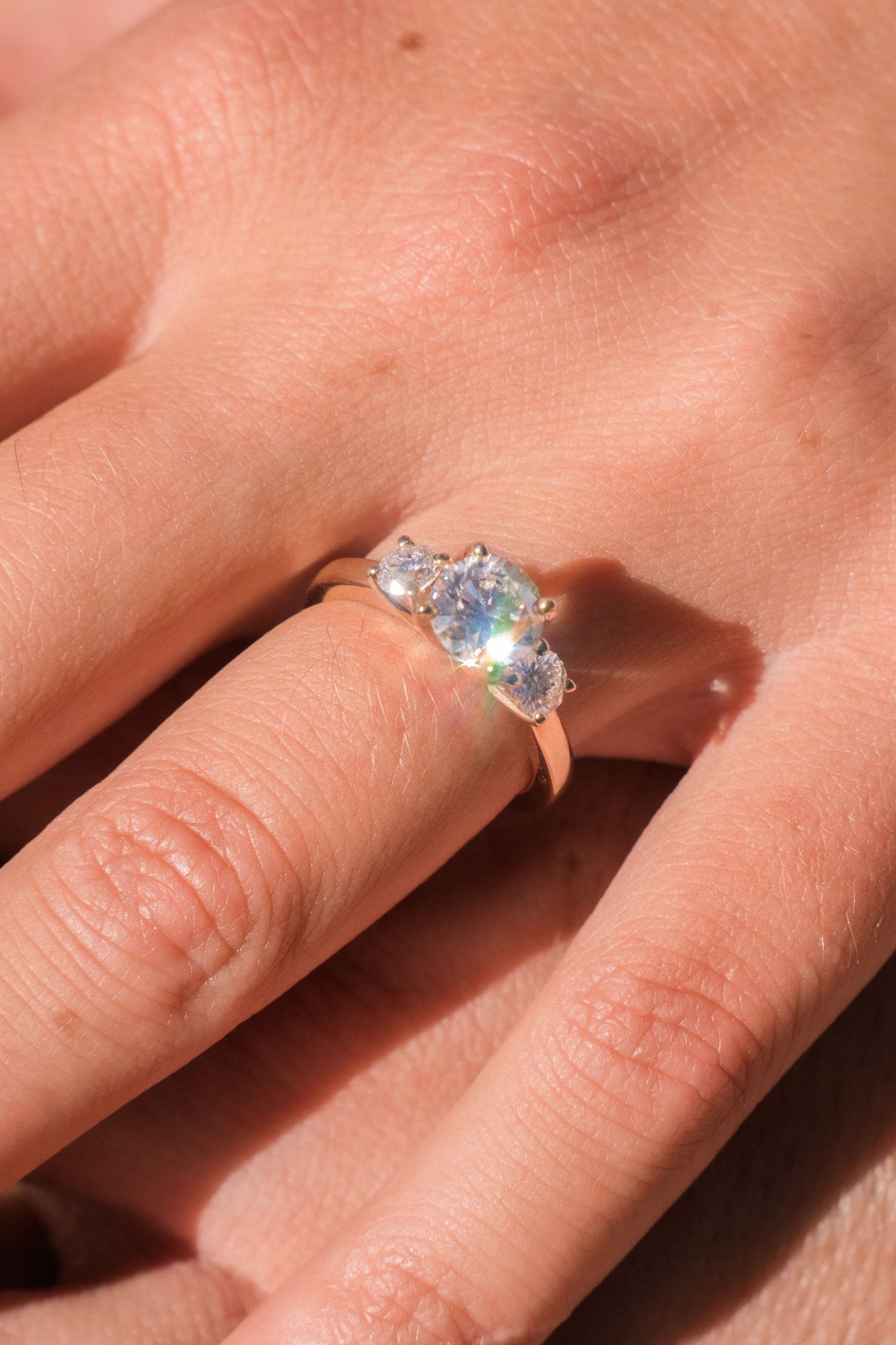 Close-up shot of a sparkling engagement ring on a hand against a pink background.