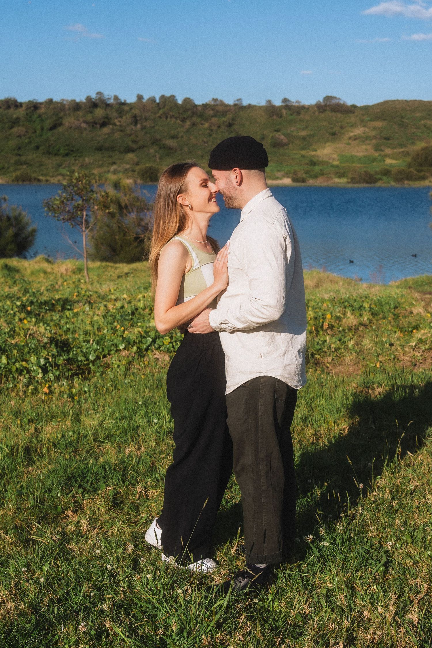 A romantic moment between two people in a grassy field overlooking a serene lake.