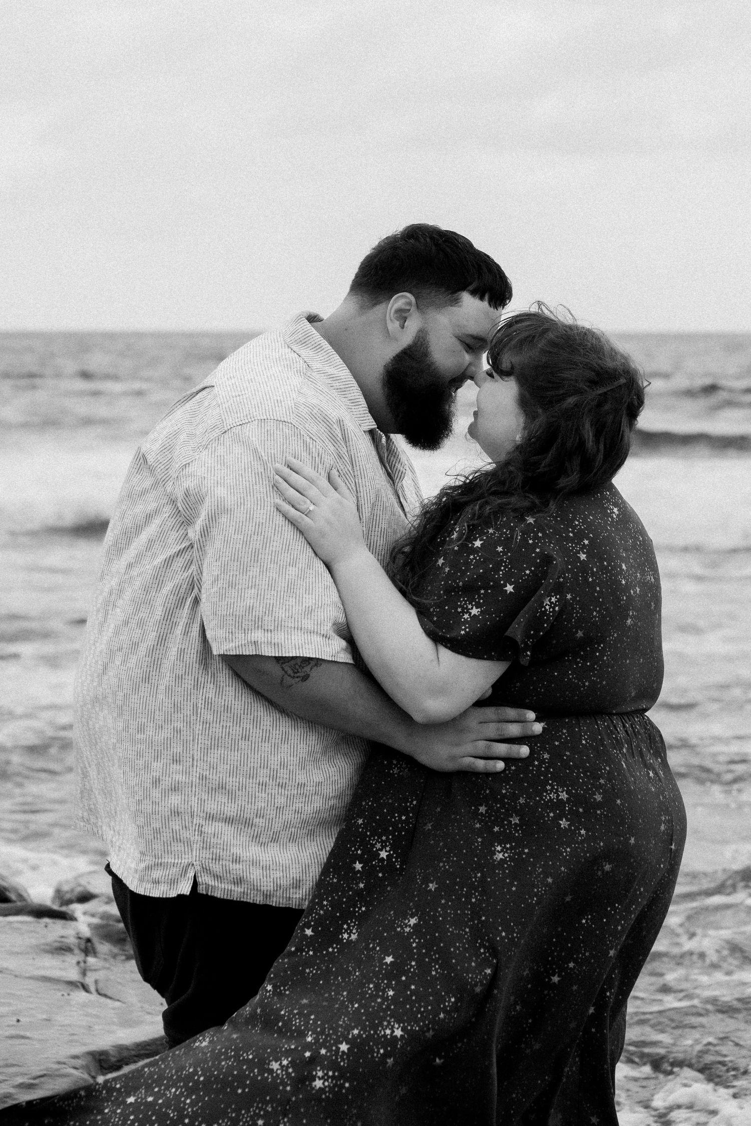 Black and white beach portrait capturing an intimate embrace against ocean waves.