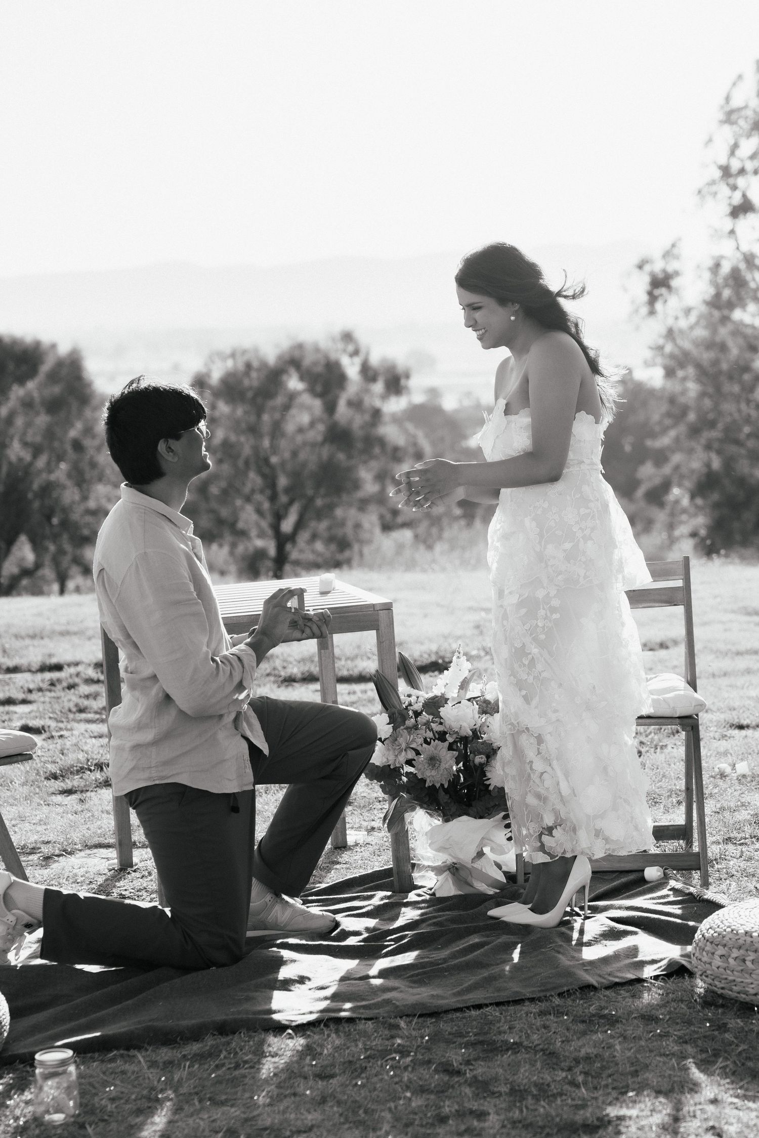 A romantic outdoor proposal scene with flowers and a picnic blanket.