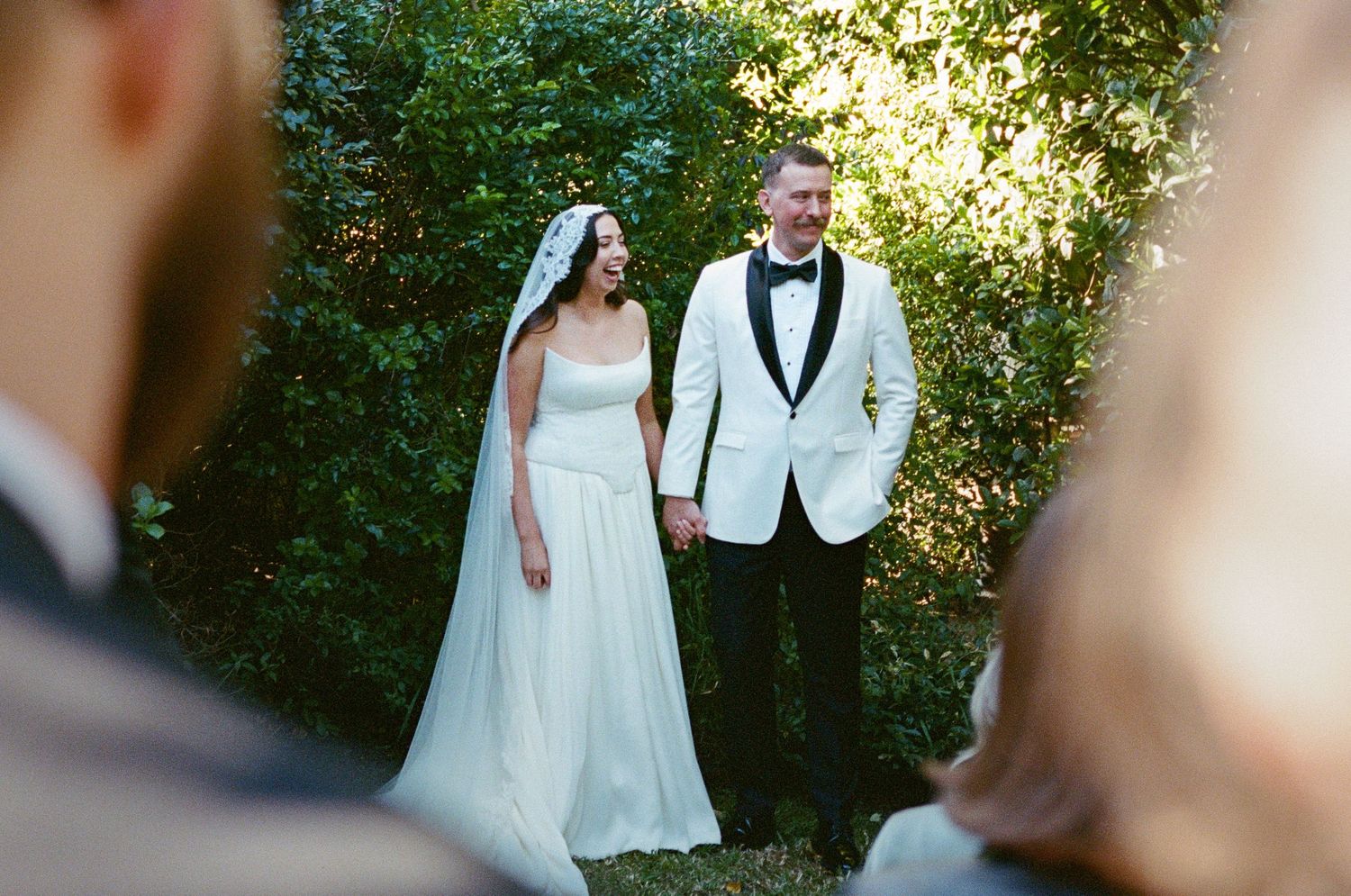 Wedding couple walking through sunlit garden path in formal attire.