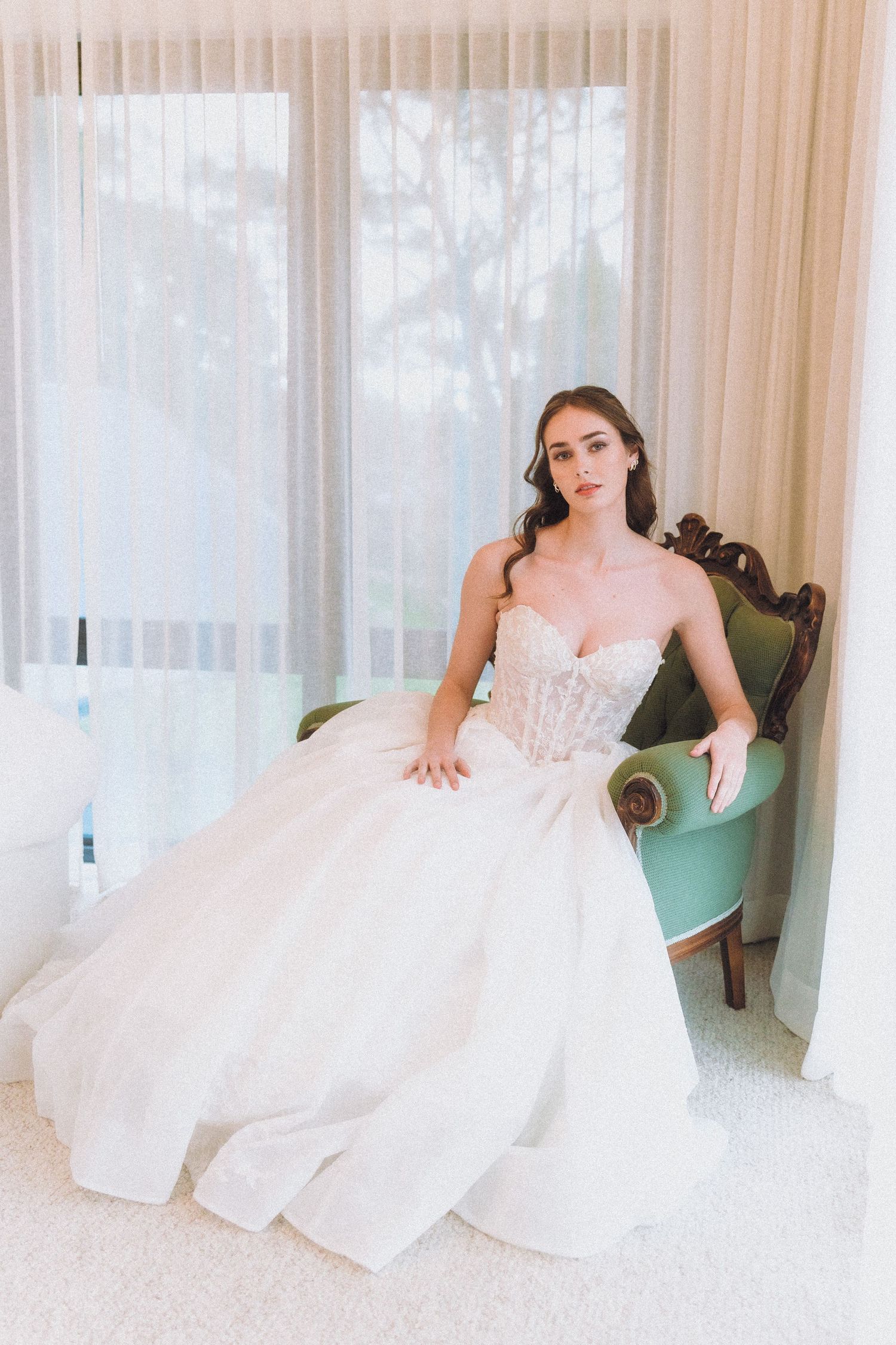 Elegant bridal portrait featuring a white ball gown and vintage-style chair against sheer curtains.