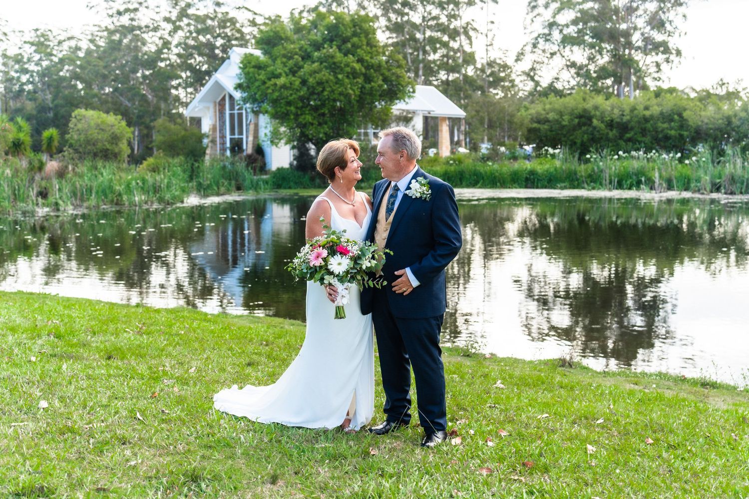 Wedding couple sitting on grass by lakeside during outdoor celebration.