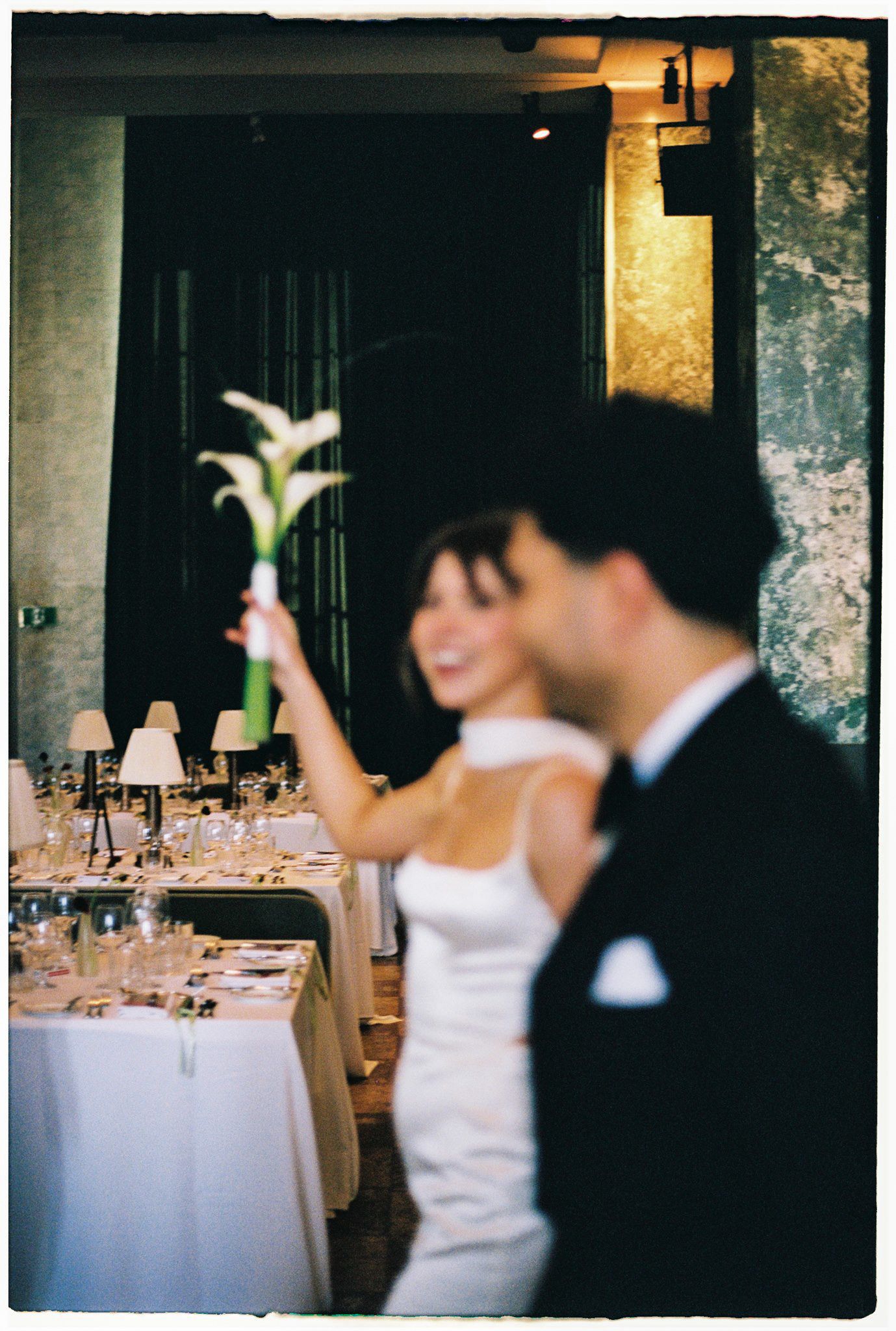 Bride in white dress raises a calla lily while standing beside groom in tuxedo at elegant wedding reception dinner.