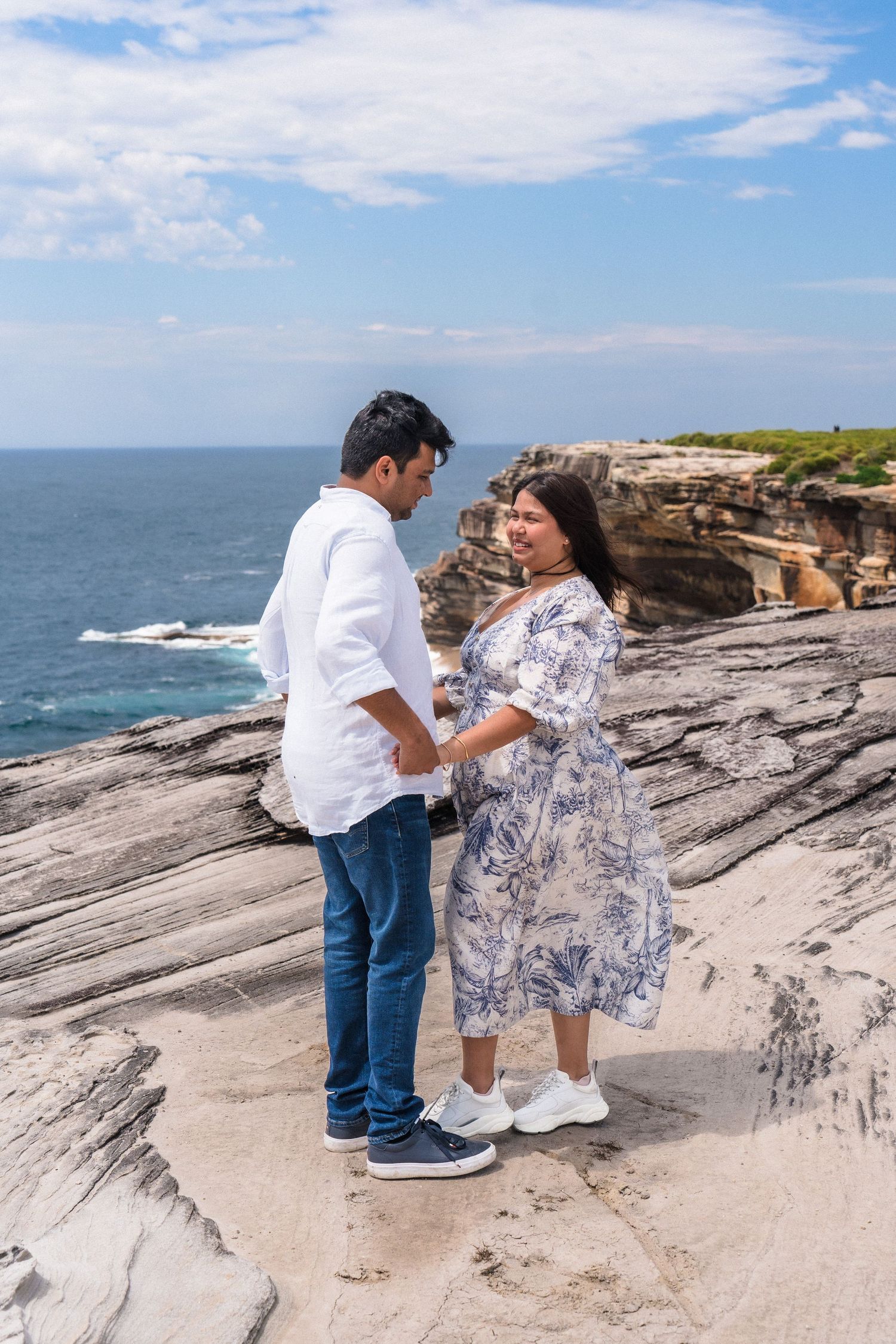 Two people share a romantic moment on rugged oceanside cliffs under blue skies.