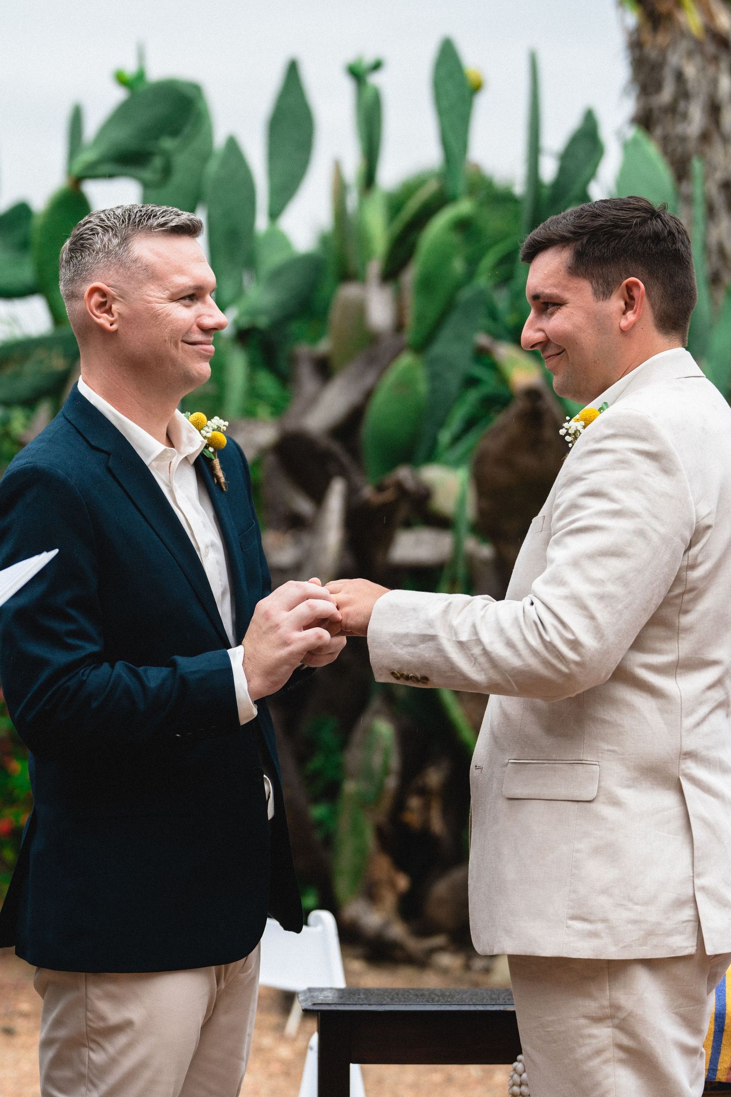 Two grooms exchanging rings during an outdoor wedding ceremony with cactus backdrop.