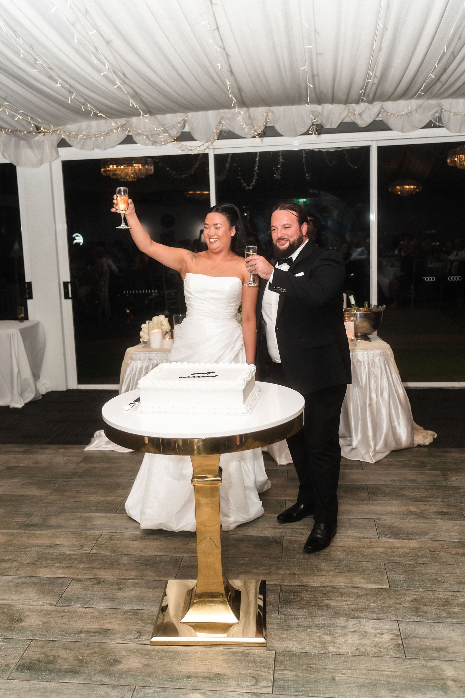 Wedding couple toasts with champagne beside their cake in decorated reception venue.