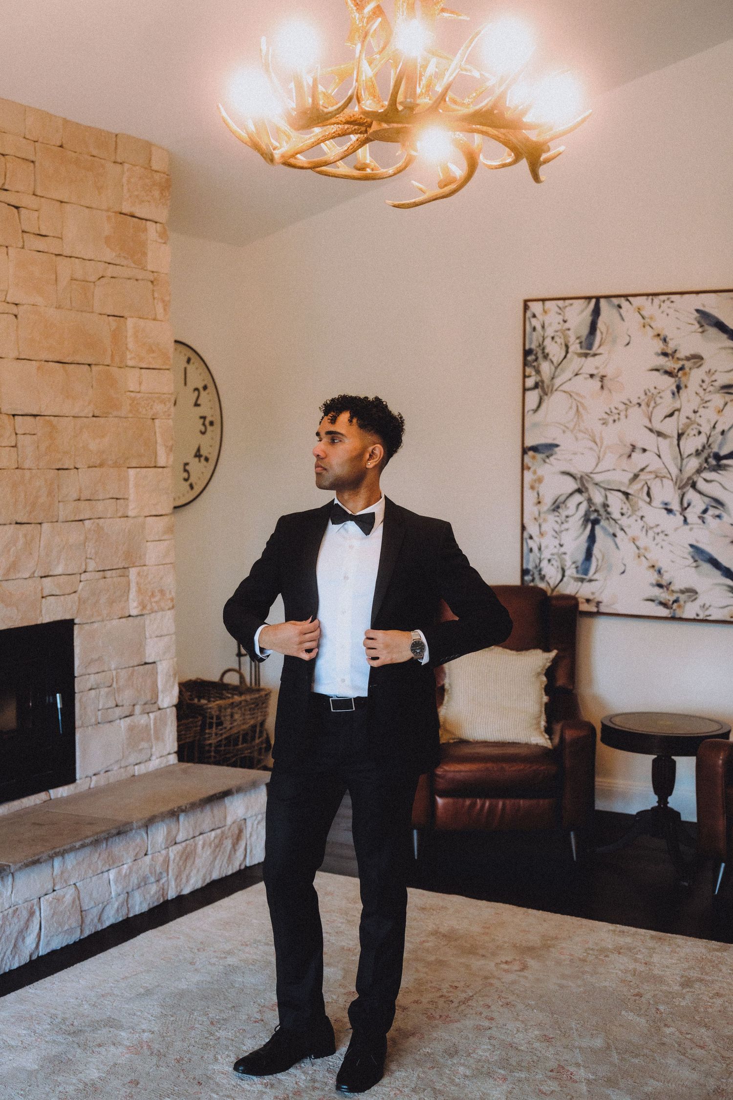 Elegant man in tuxedo adjusting his jacket while standing in a modern room with antler chandelier.