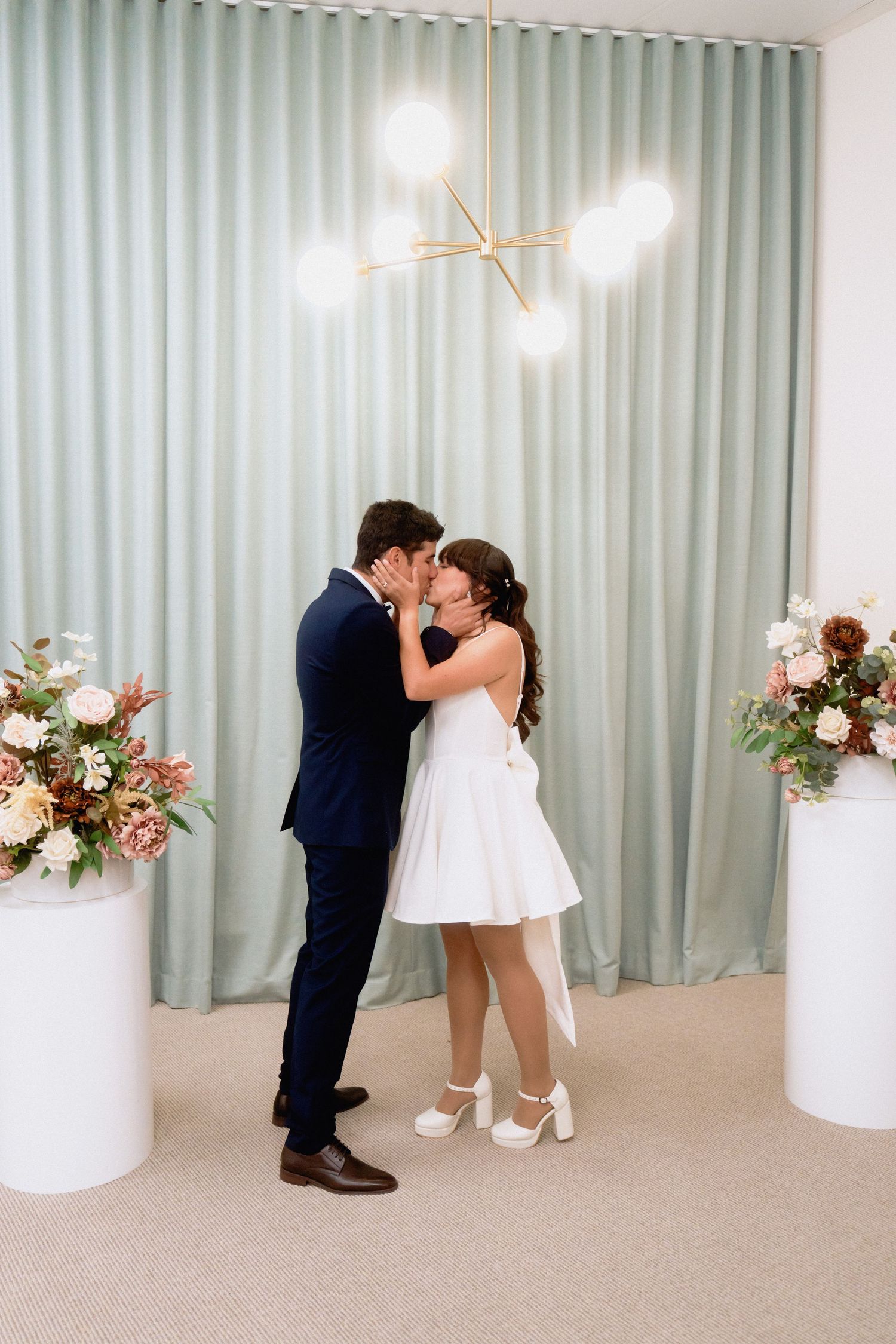 Couple sharing intimate moment in modern wedding venue with sage curtains and floral arrangements.