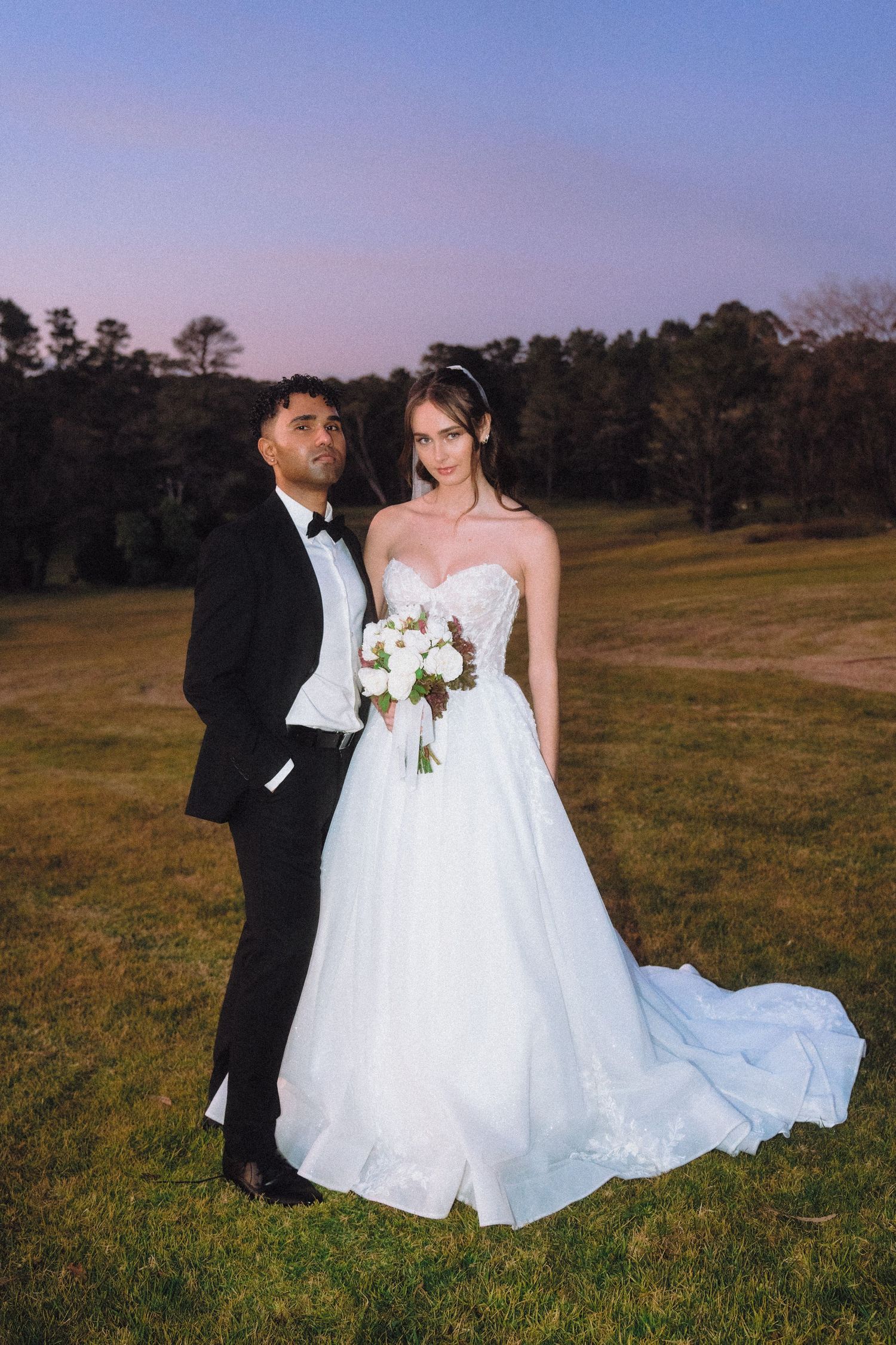 Wedding couple poses in open field with flowing white gown and classic tuxedo.