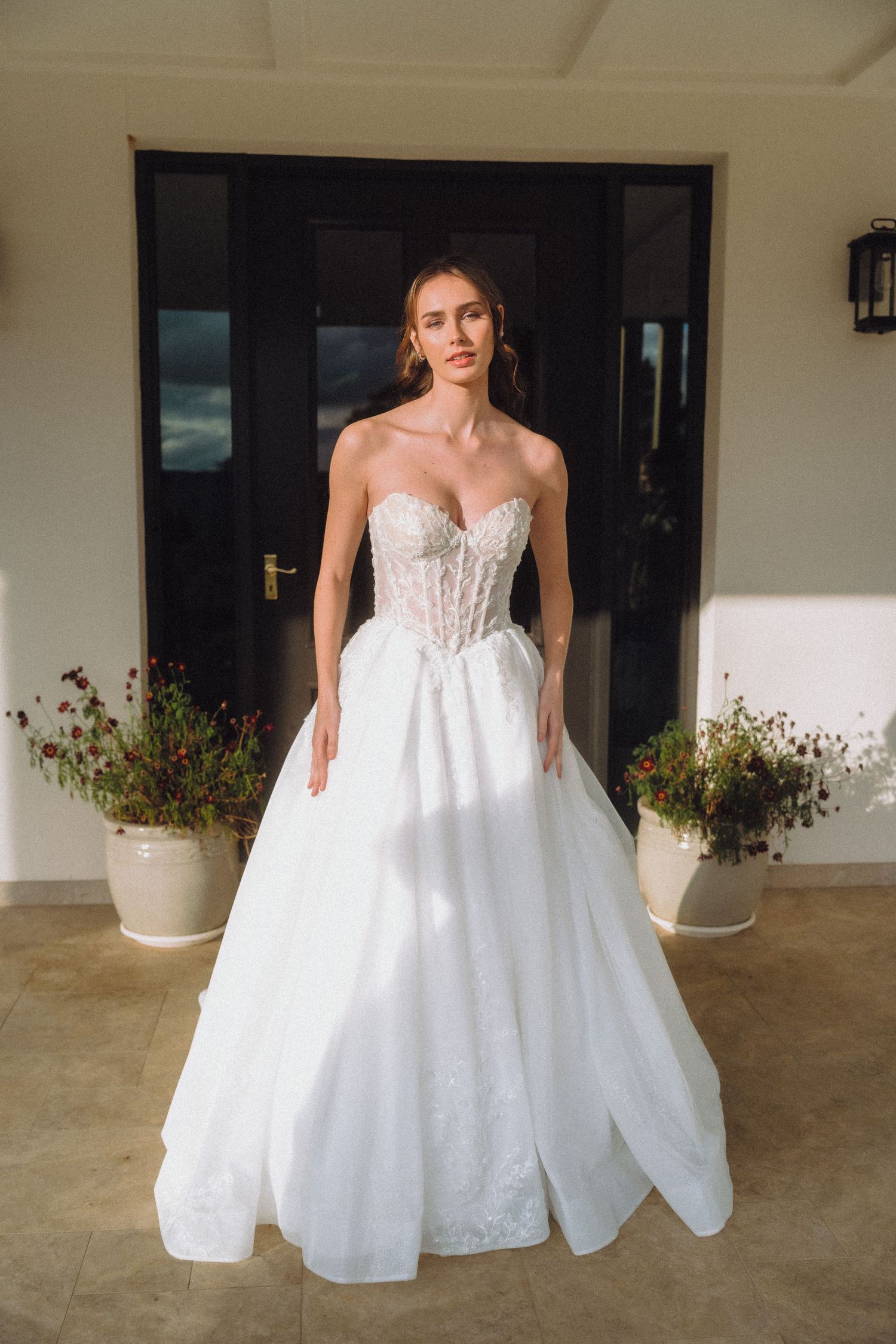 Bride in white ballgown poses on porch with potted flowers and classic architecture.