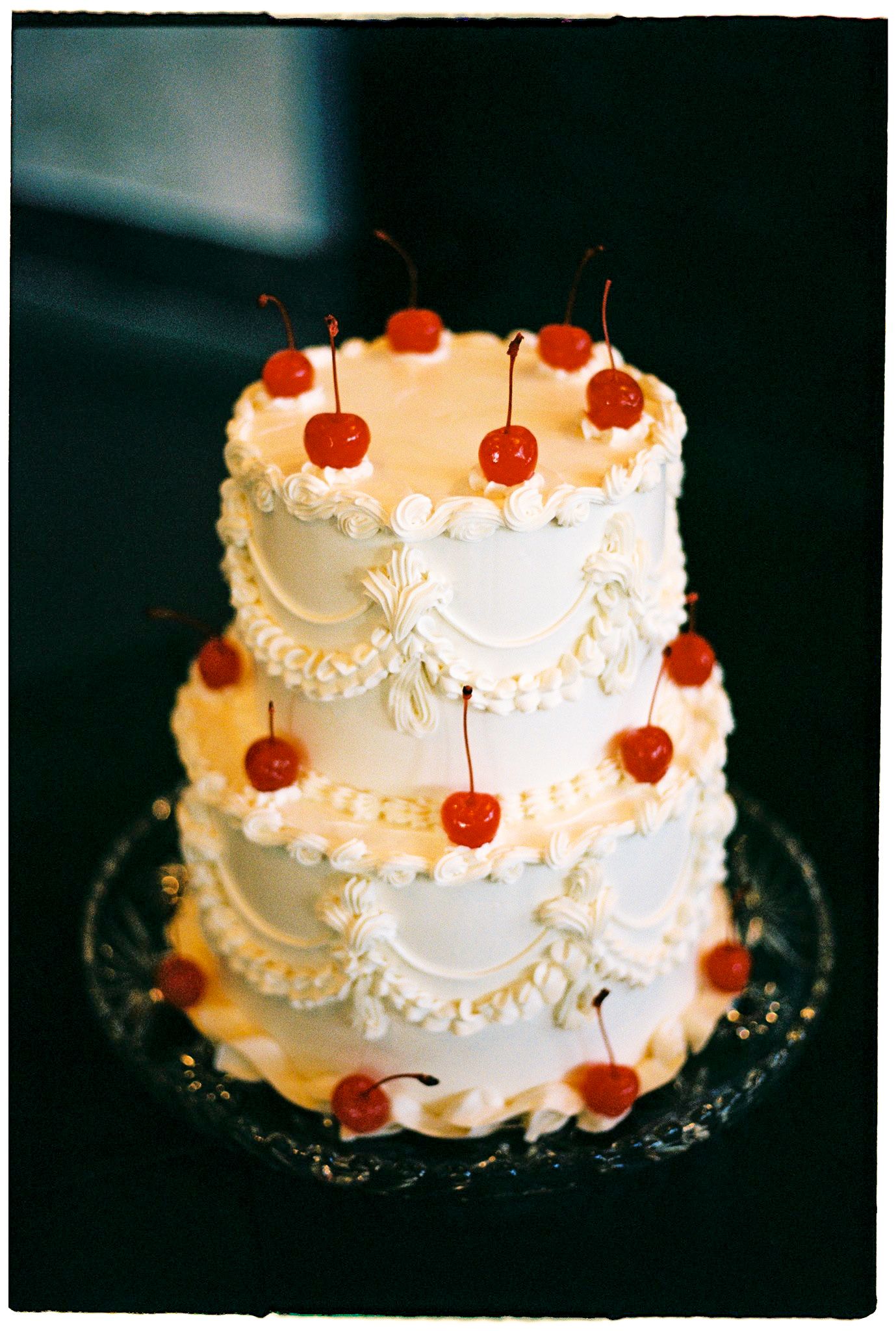 Three-tier white wedding cake decorated with maraschino cherries and intricate piped frosting details on a glass stand.