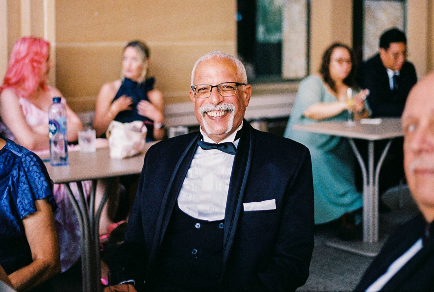 Smiling elderly man in tuxedo with bow tie at formal event, guests seated at tables in background.
