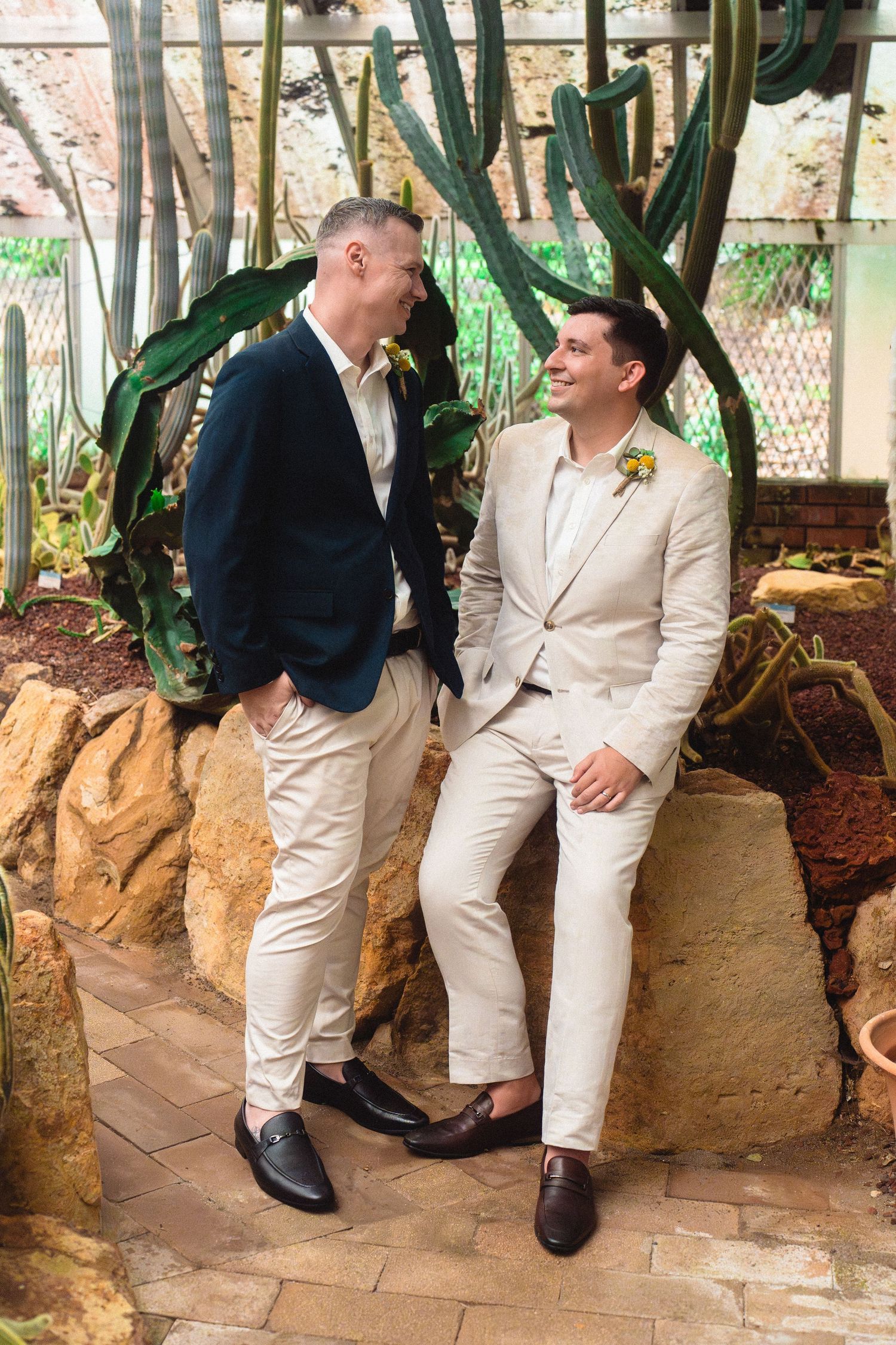 Two grooms in suits smiling at each other among cacti on their wedding day.