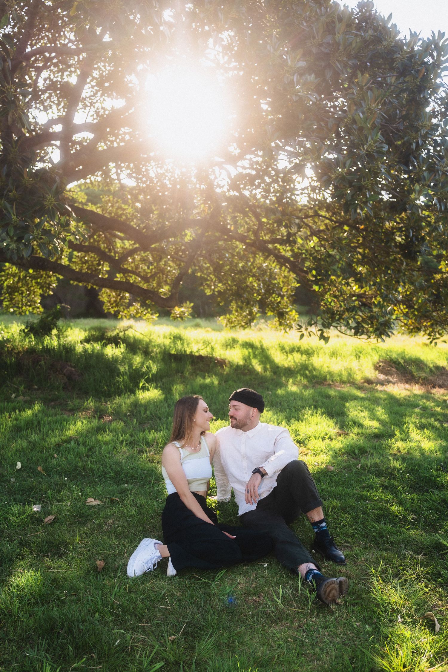 A couple shares a tender moment on grass during golden hour with sunlight streaming through trees.