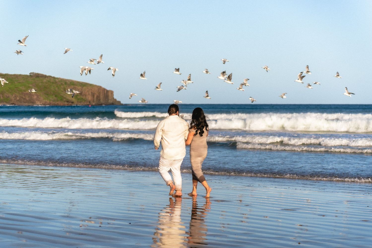 A couple strolls along the shoreline with seabirds flying above.