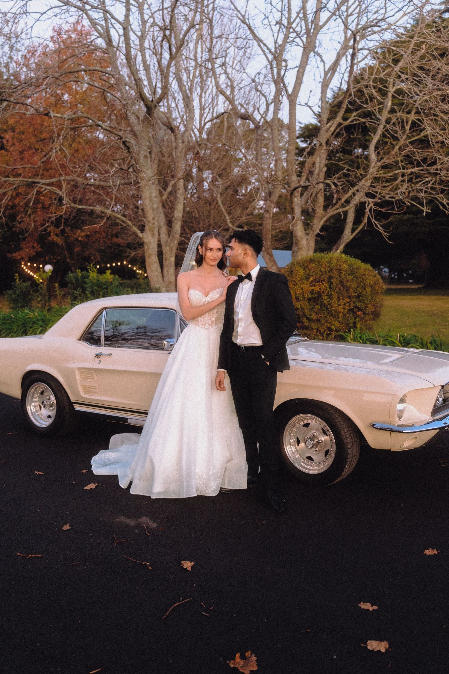 Wedding couple poses romantically by vintage car during autumn sunset.