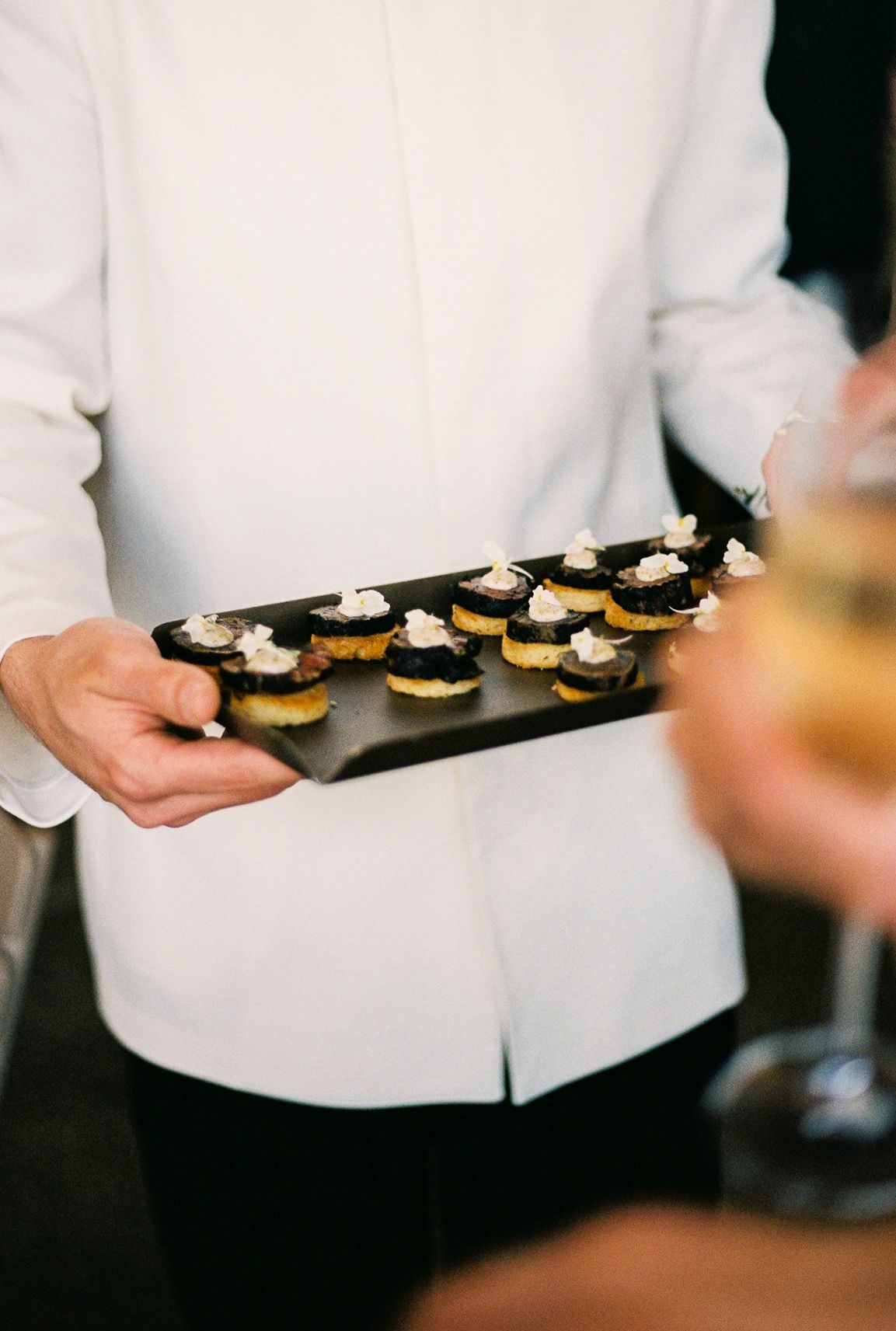 Server holding a black slate tray of elegant caviar and cream canapes at an upscale catered event.