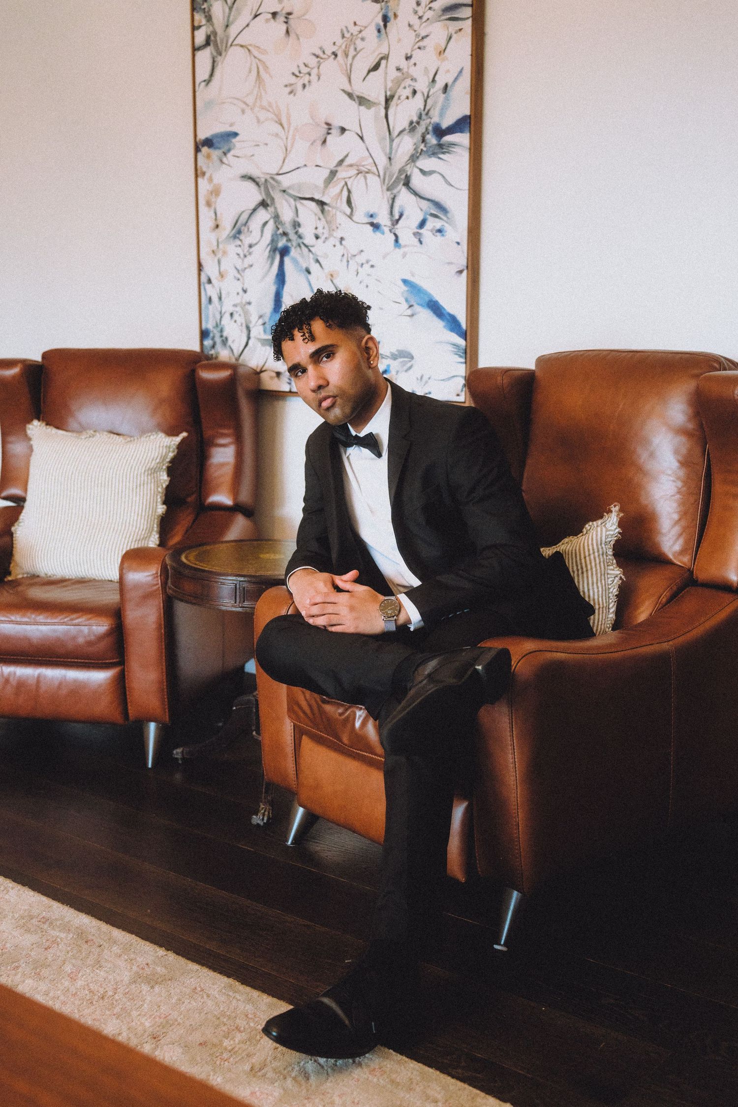 Stylish man in formal attire seated on brown leather chair with artistic wall decor behind.