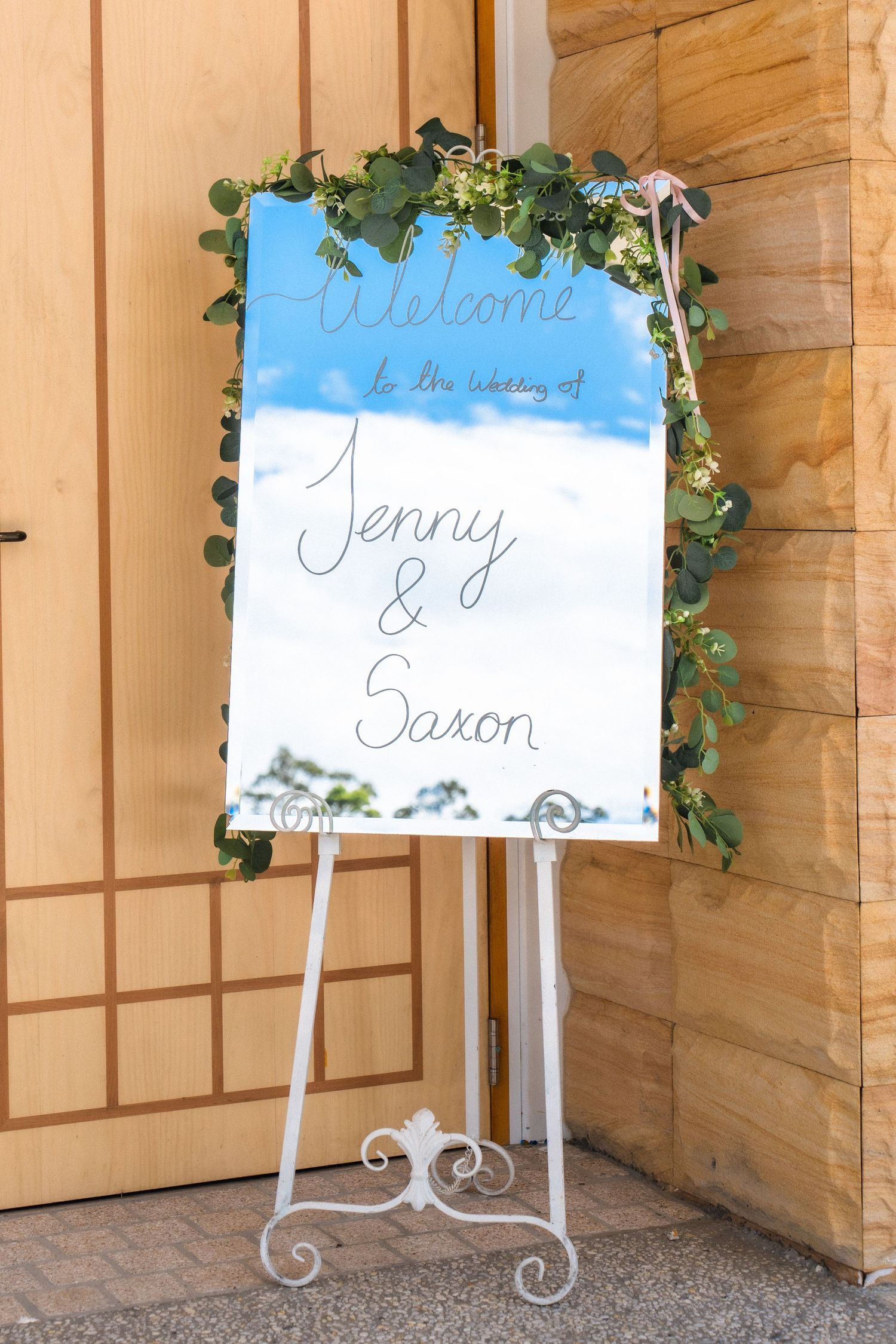 Decorative wedding welcome sign with greenery accents against wooden panels.
