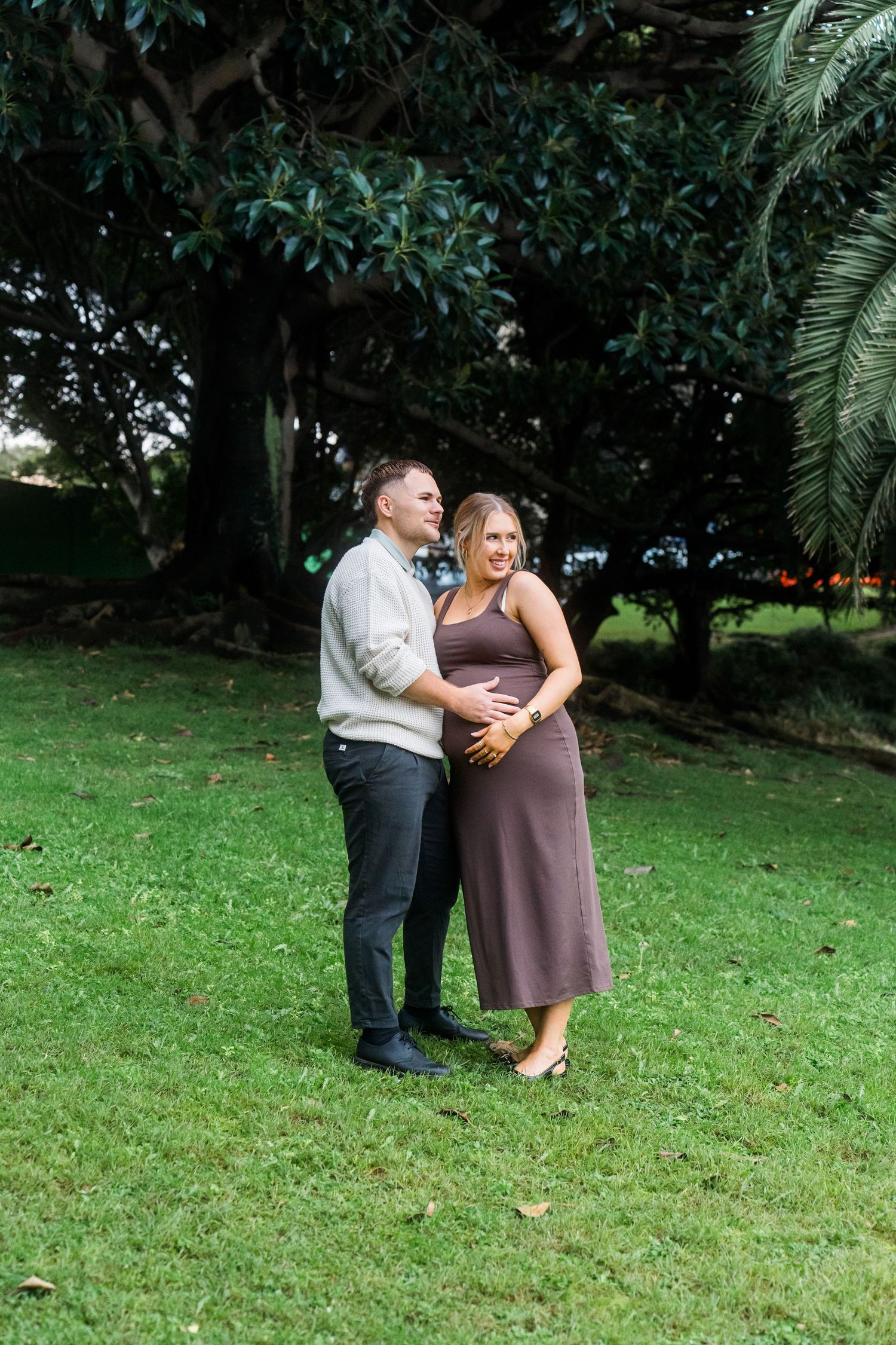 A couple shares an intimate moment while standing on lush green grass with palm trees.