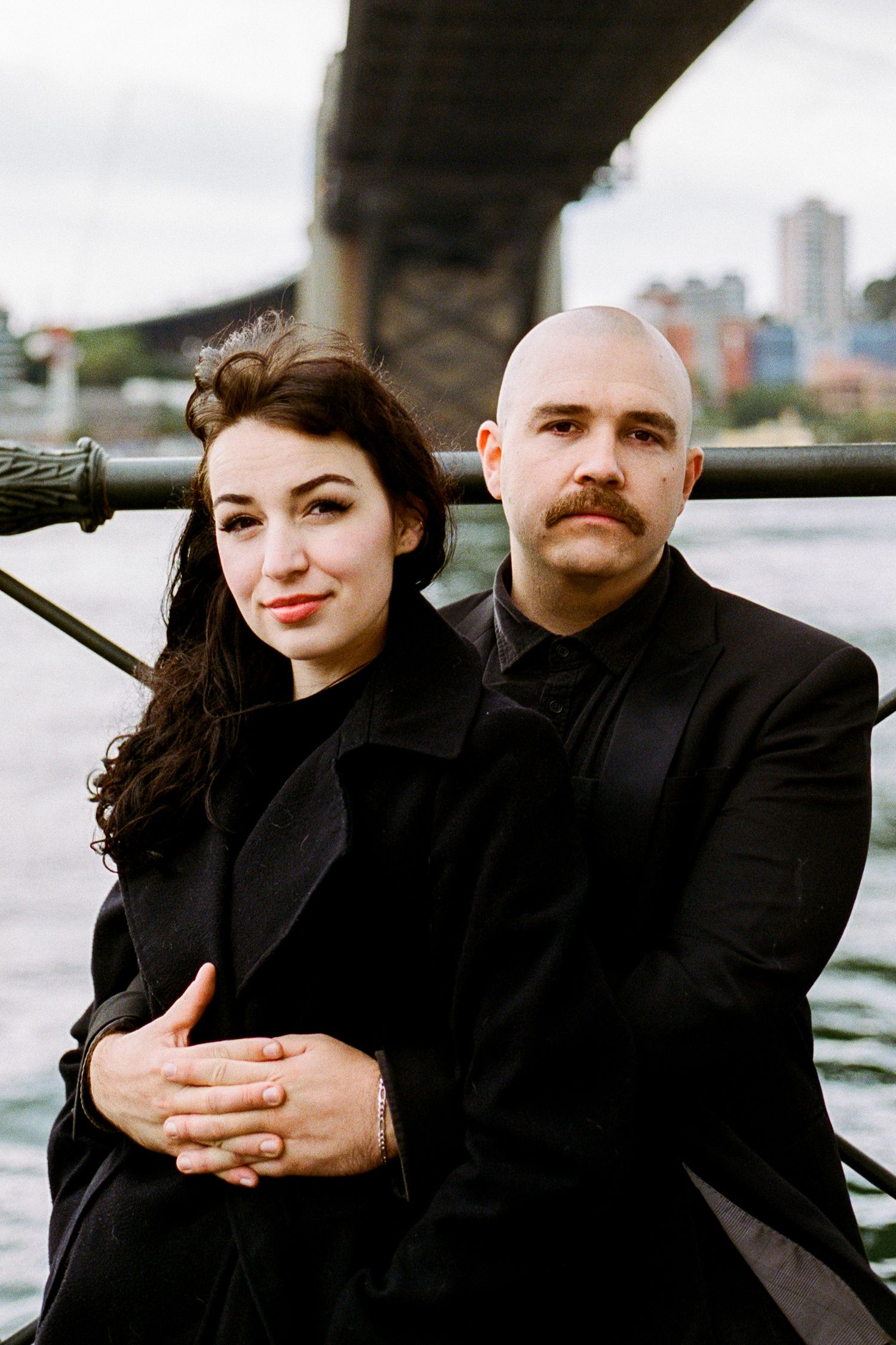 A couple in black attire poses intimately against a waterfront cityscape backdrop.