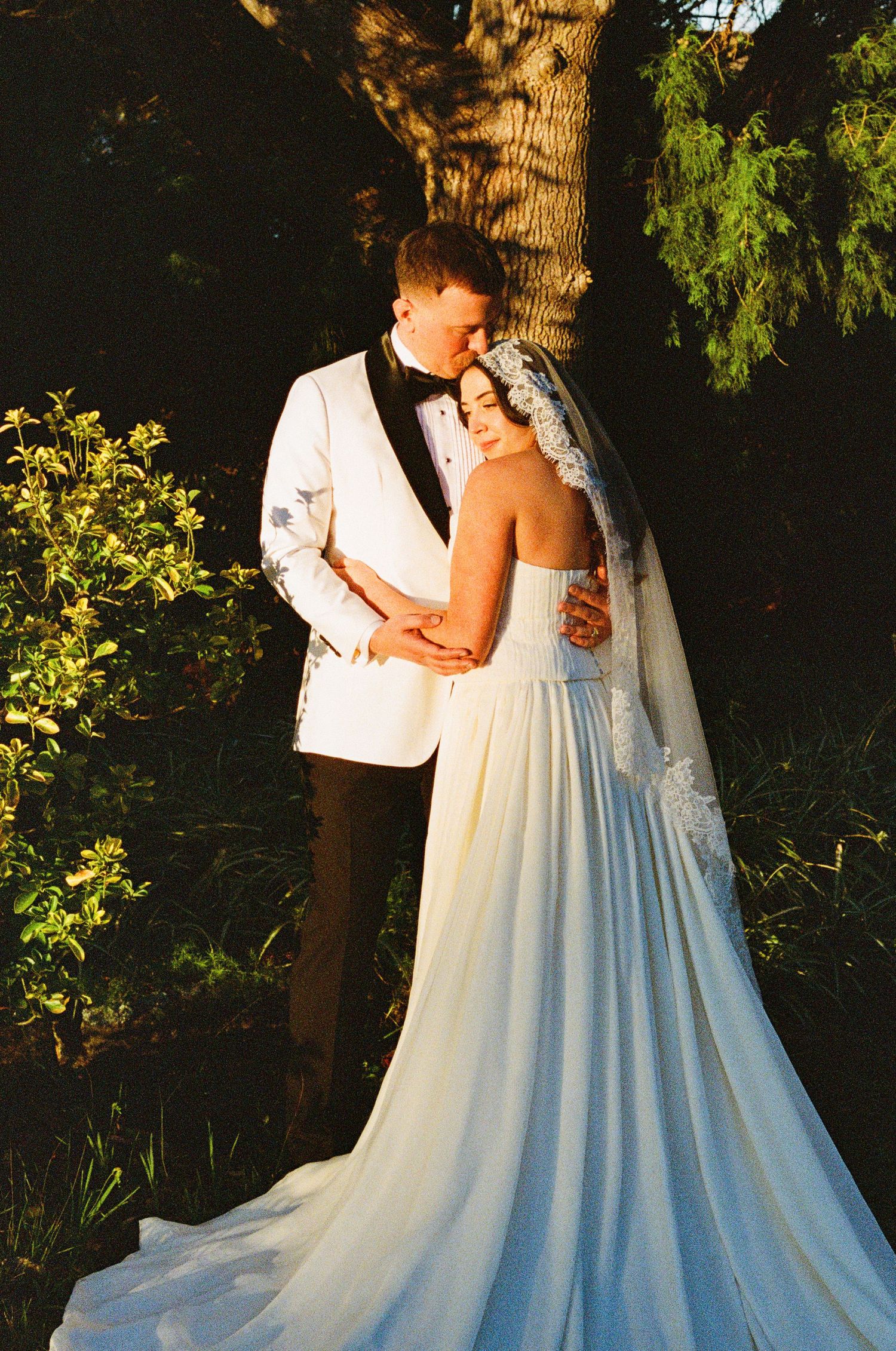 Bride in flowing white gown embraces partner during sunset wedding photoshoot.