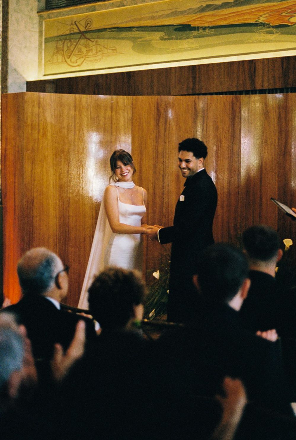 Bride and groom smiling and holding hands during wedding ceremony in elegant wood-paneled venue.