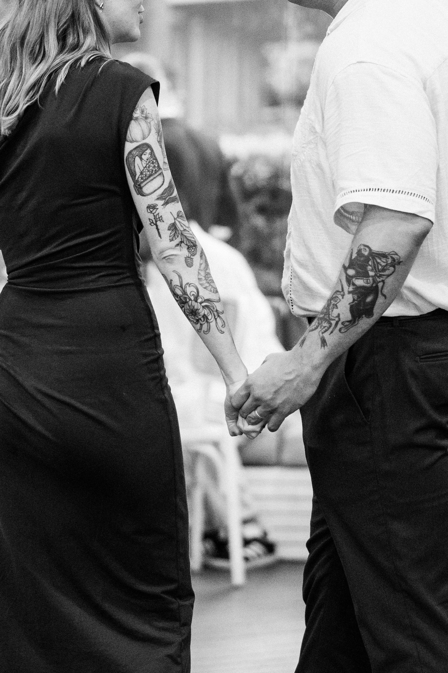 Black and white photo of a tattooed couple holding hands, woman in black dress, man in white shirt.