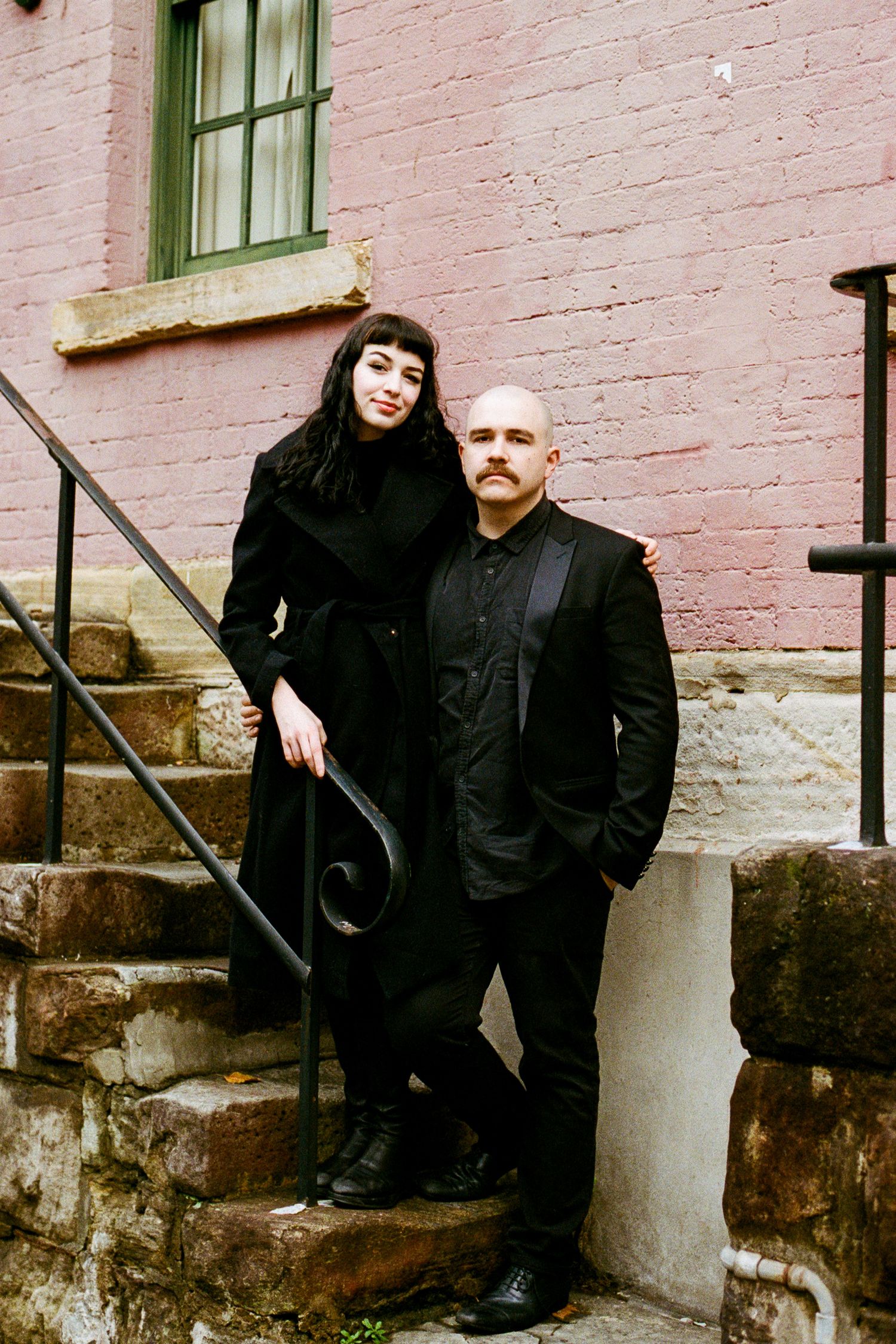 A couple in dark clothing poses against a pink brick wall with green window frames.