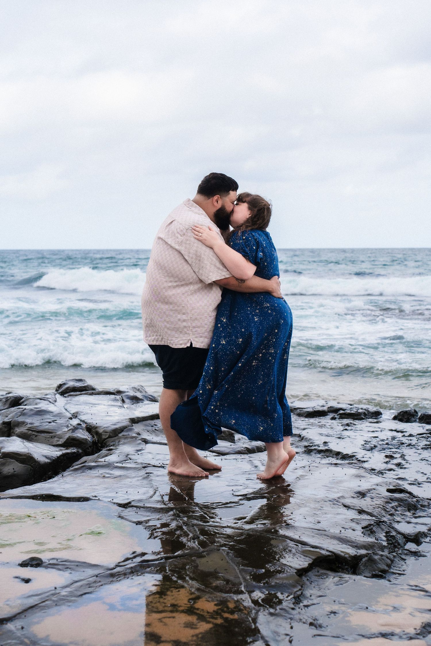 Couple embracing on rocky coastal shoreline with crashing waves in the background.