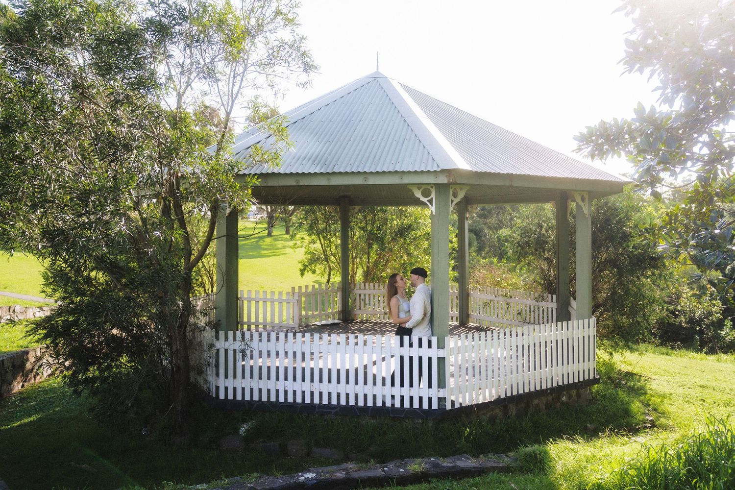 White gazebo with picket fence surrounded by lush greenery in sunlight.