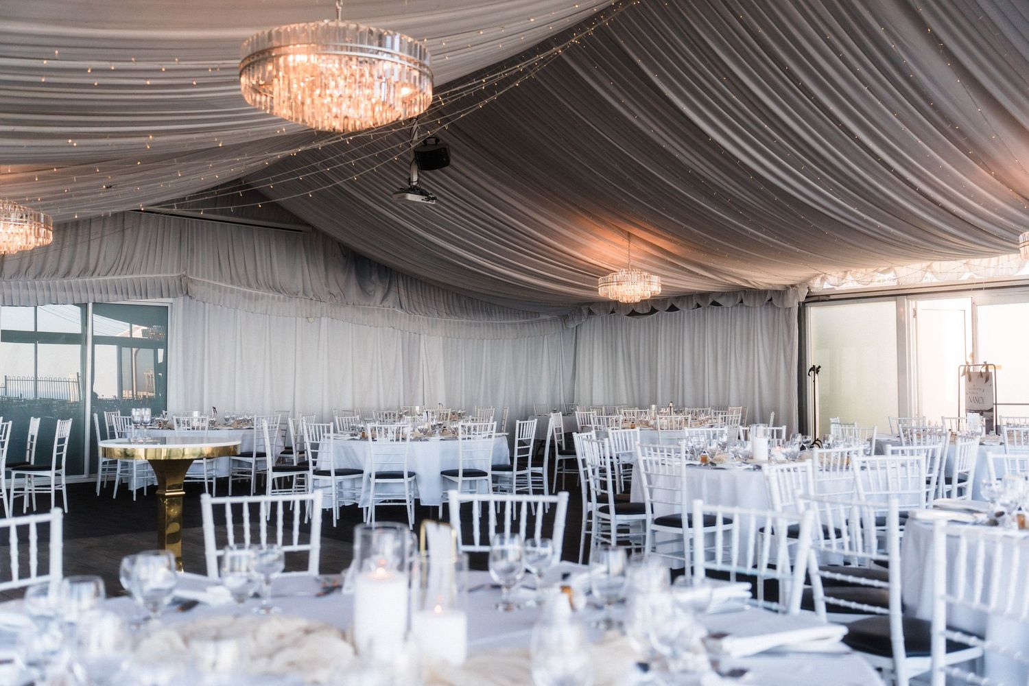Elegant wedding reception venue with white draped ceiling, crystal chandeliers and chiavari chairs.