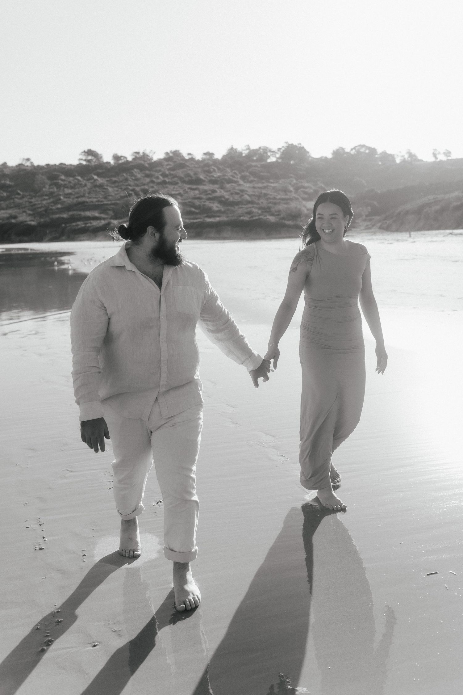 Two people in white clothing walk together on a beach casting long shadows.