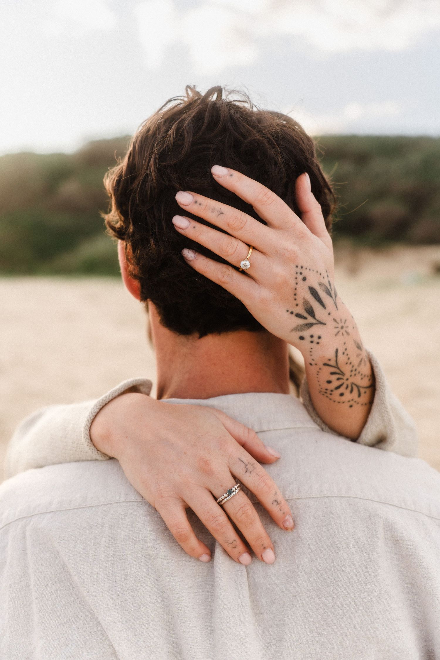Close-up of hands with henna tattoo and ring embracing on beach at sunset.