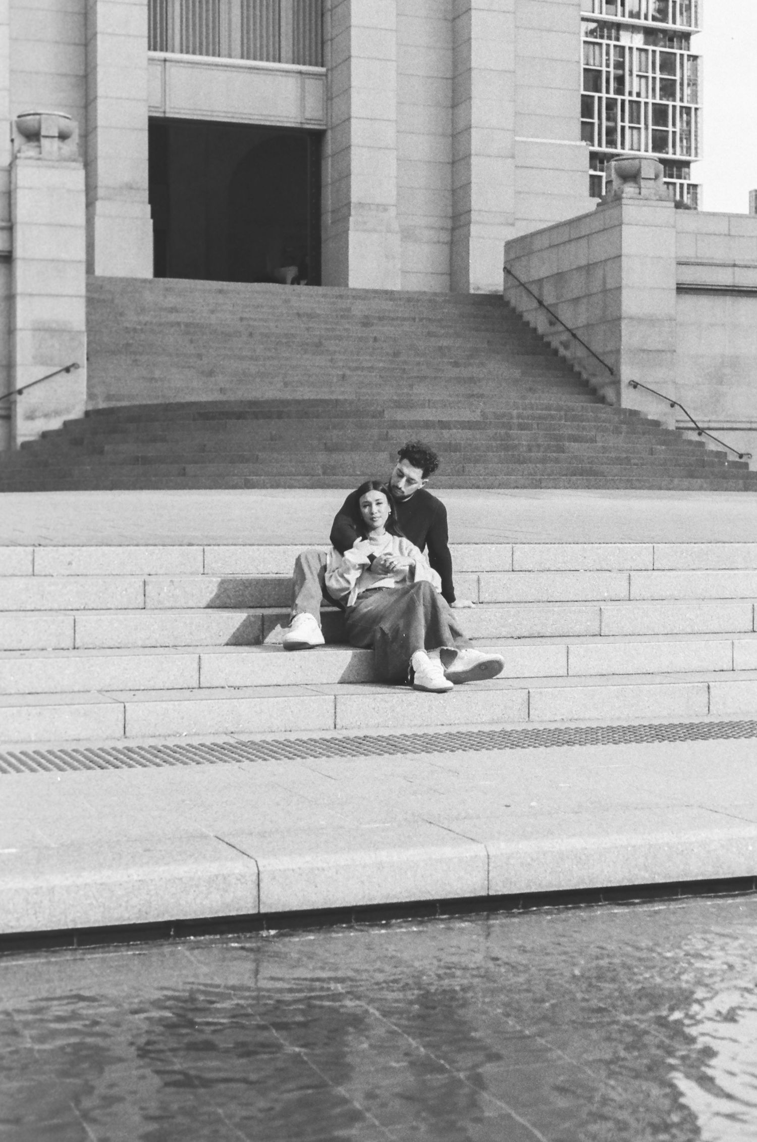 A couple sits together on concrete steps near a reflecting pool.