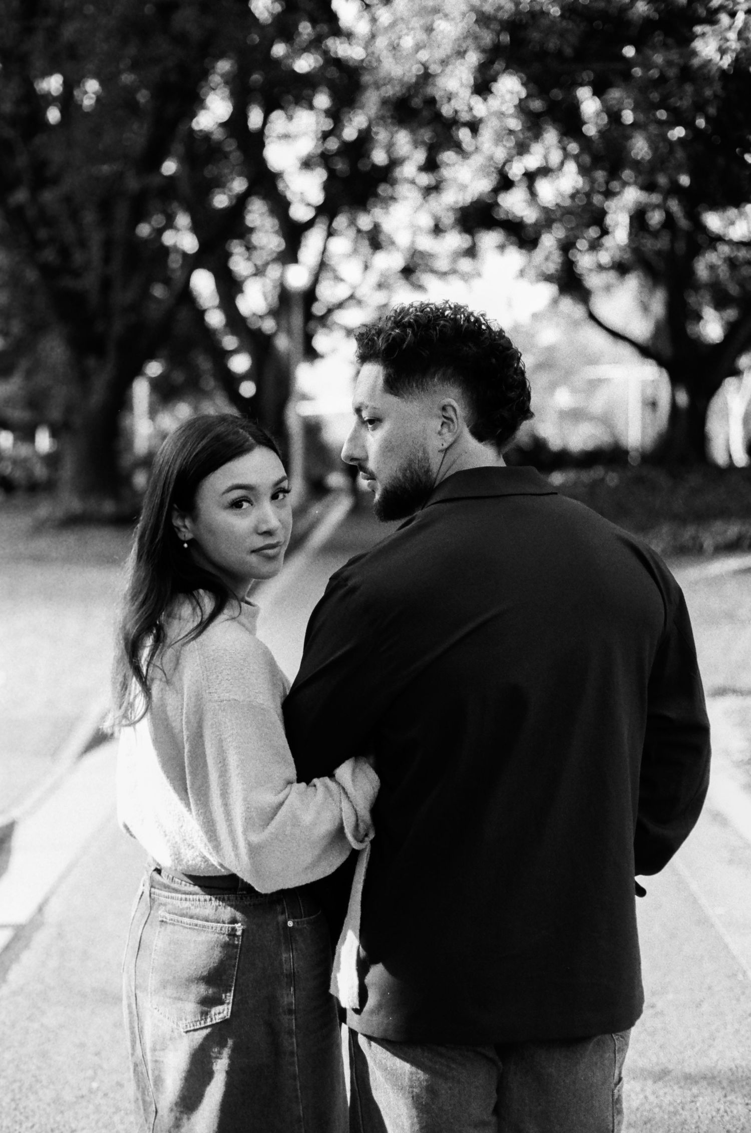 A couple stands close together on a tree-lined path in black and white.