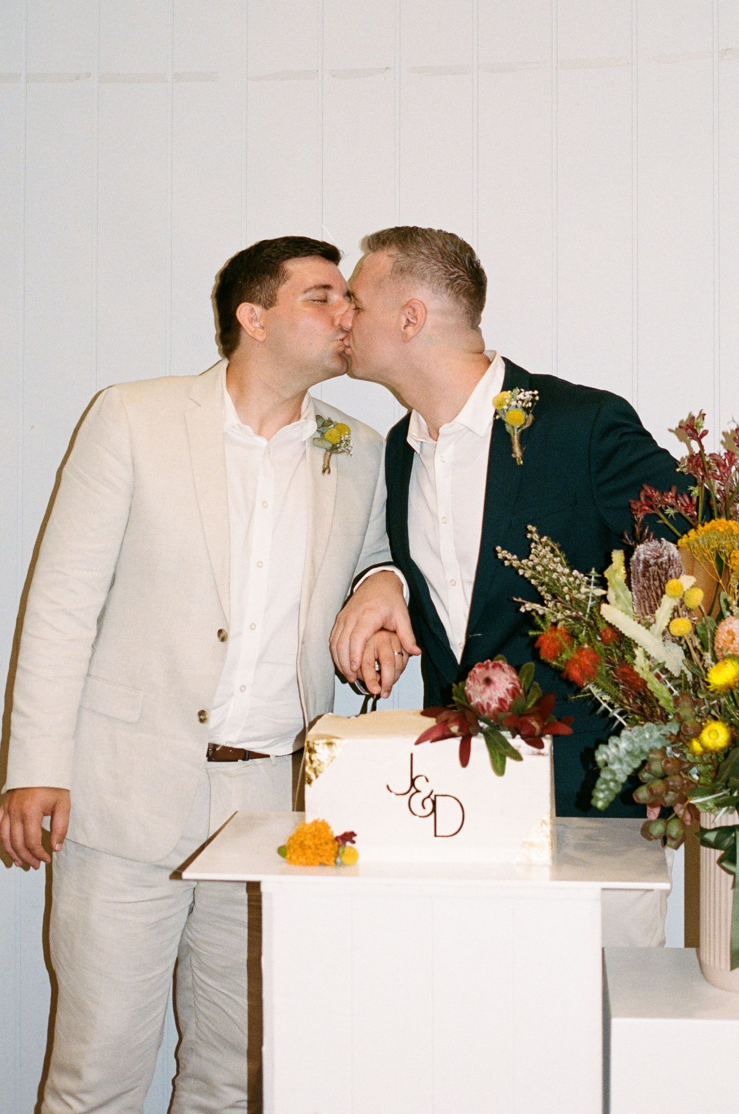 Two grooms share a kiss while cutting their wedding cake, adorned with initials J&D, surrounded by colorful floral arrangements.